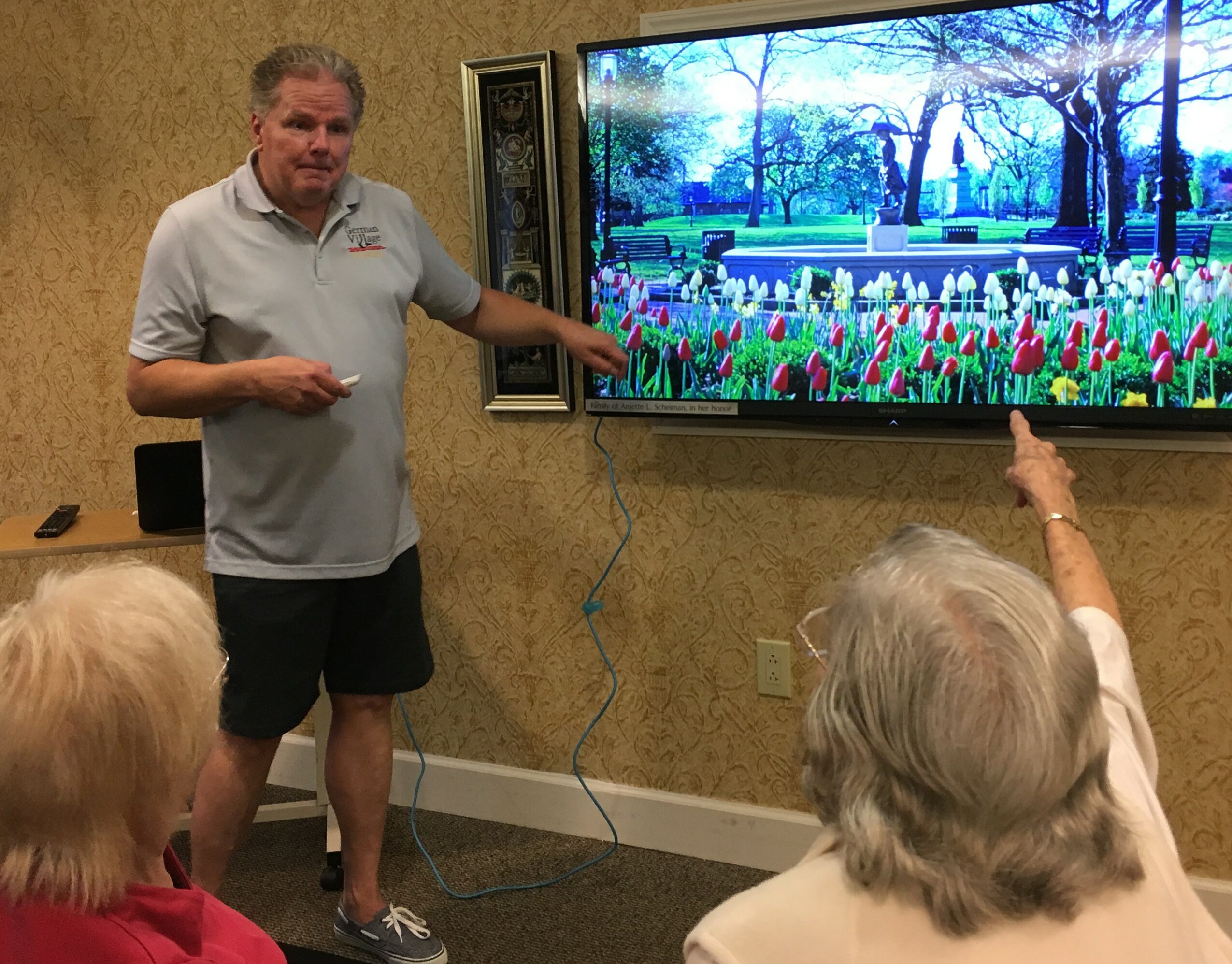 Man in white shirt and shorts points at a large screen displaying a park scene with trees and flowers, with two elderly women in foreground, one pointing at the screen.