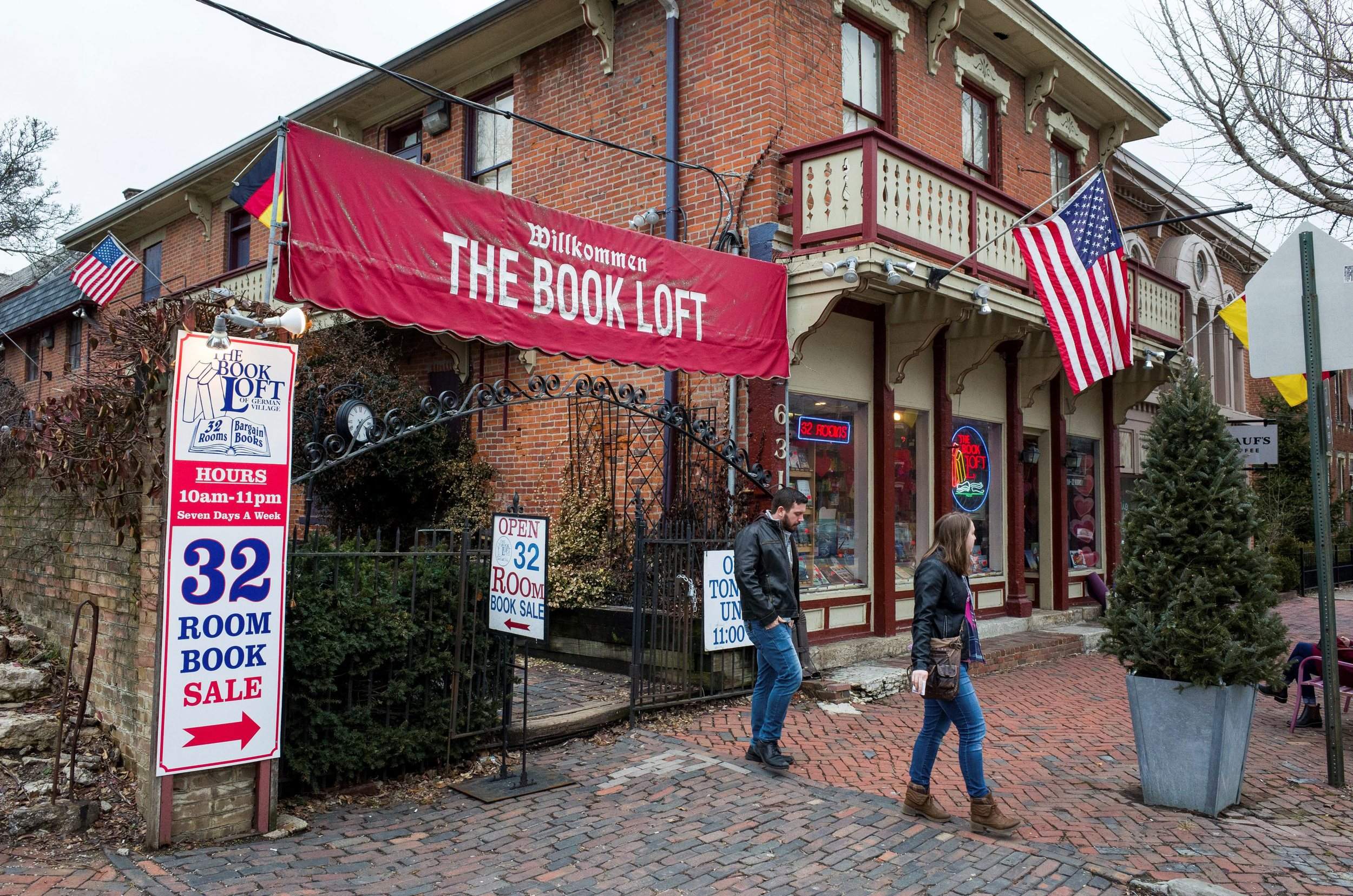 Brick building with a red banner reading 'The Book Loft' and American flags, people walking outside, signs advertising a book sale.