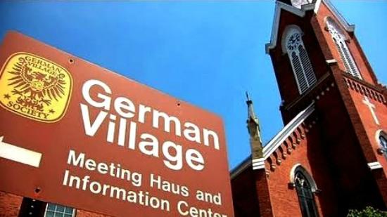 Red brick church with tall steeple against blue sky, and a sign reading 'German Village' with additional information.