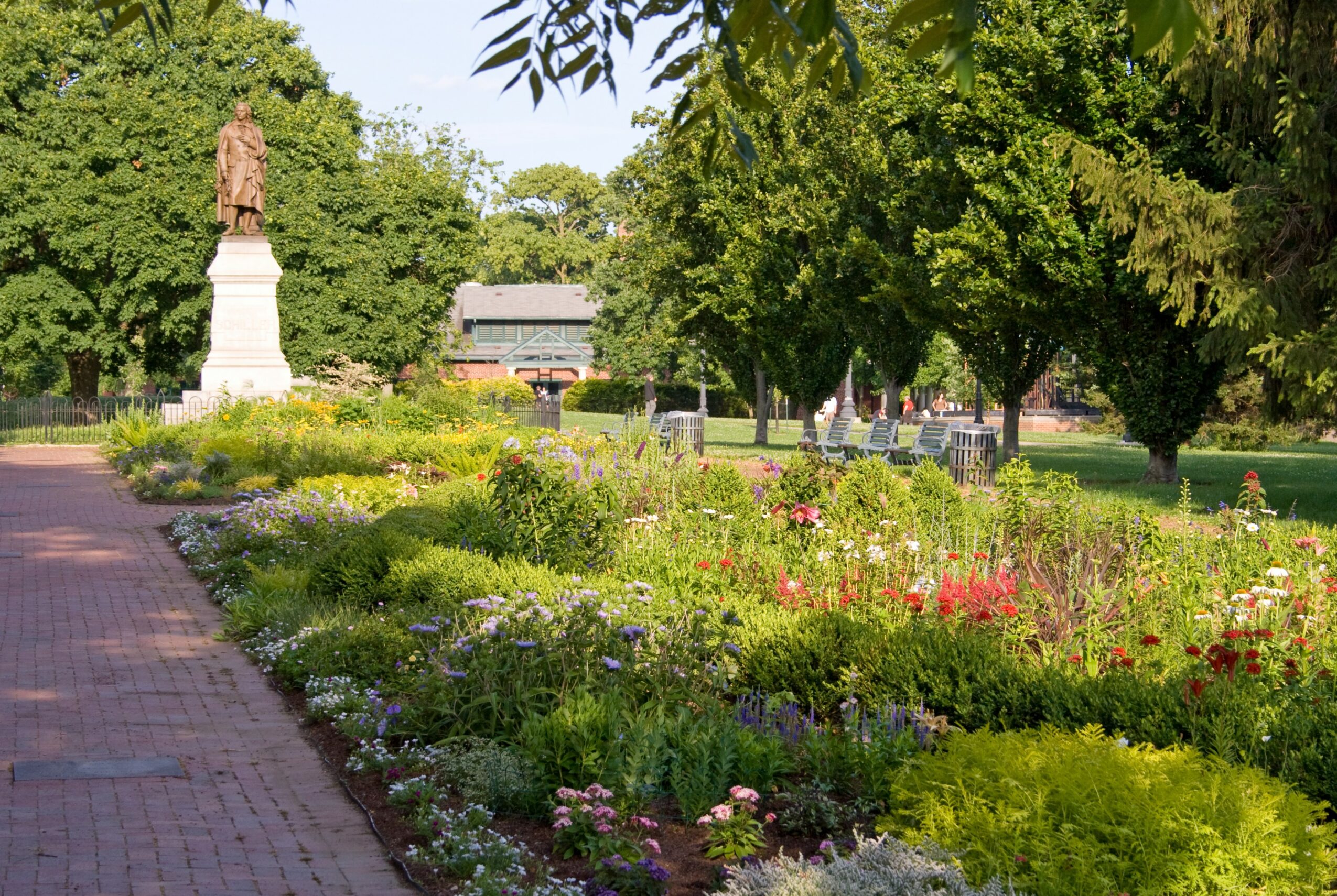 Park with flower beds, trees, a statue on a pedestal, and a pathway, under a clear sky.