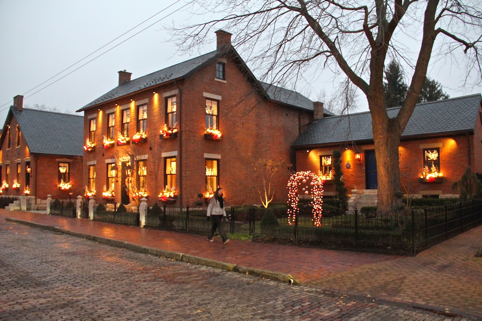 Brick building with lit windows and Christmas decorations, sidewalk, trees, and a person walking outside during evening.