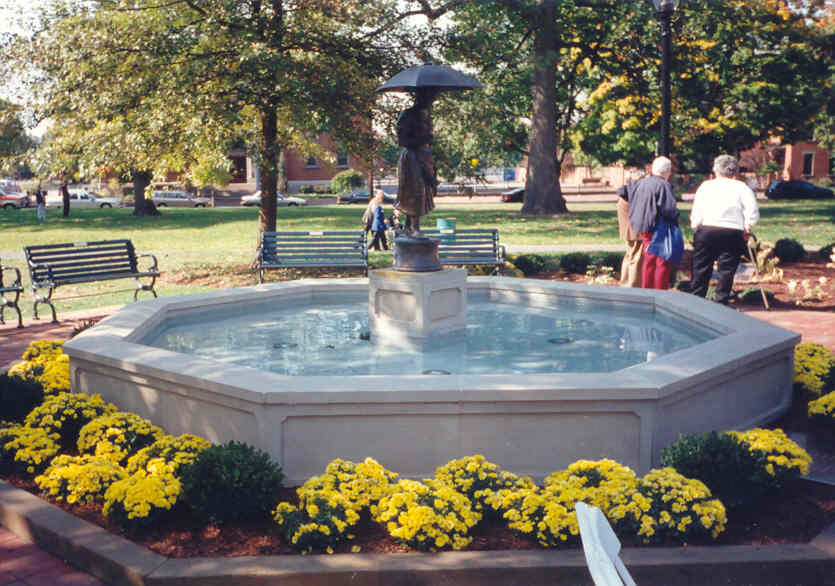 Octagonal fountain with a statue in the center, surrounded by yellow flowers and trees in a park, people walking nearby.