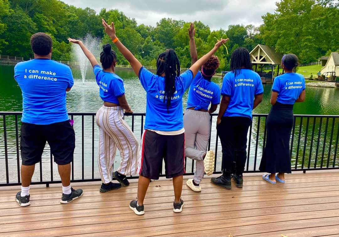 Group of six people wearing blue shirts standing on a wooden deck by a lake, with trees and houses in the background.