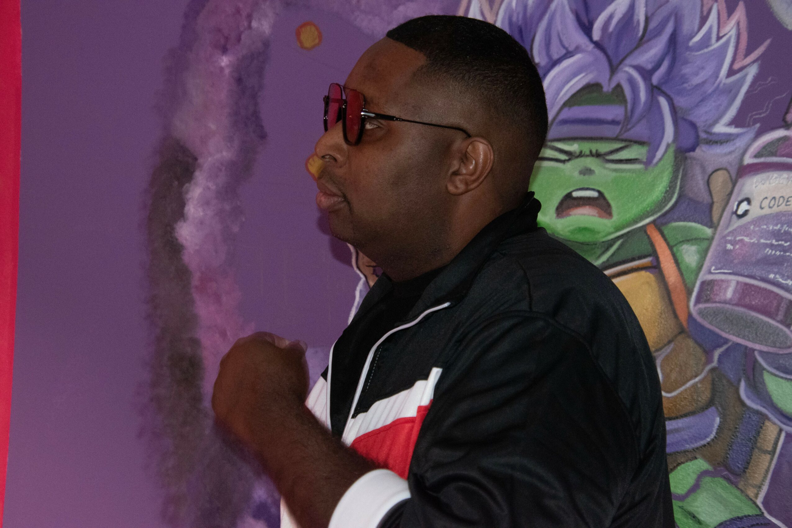 Side view of a man wearing glasses and a black jacket with red and white stripes, standing in front of a colorful mural.