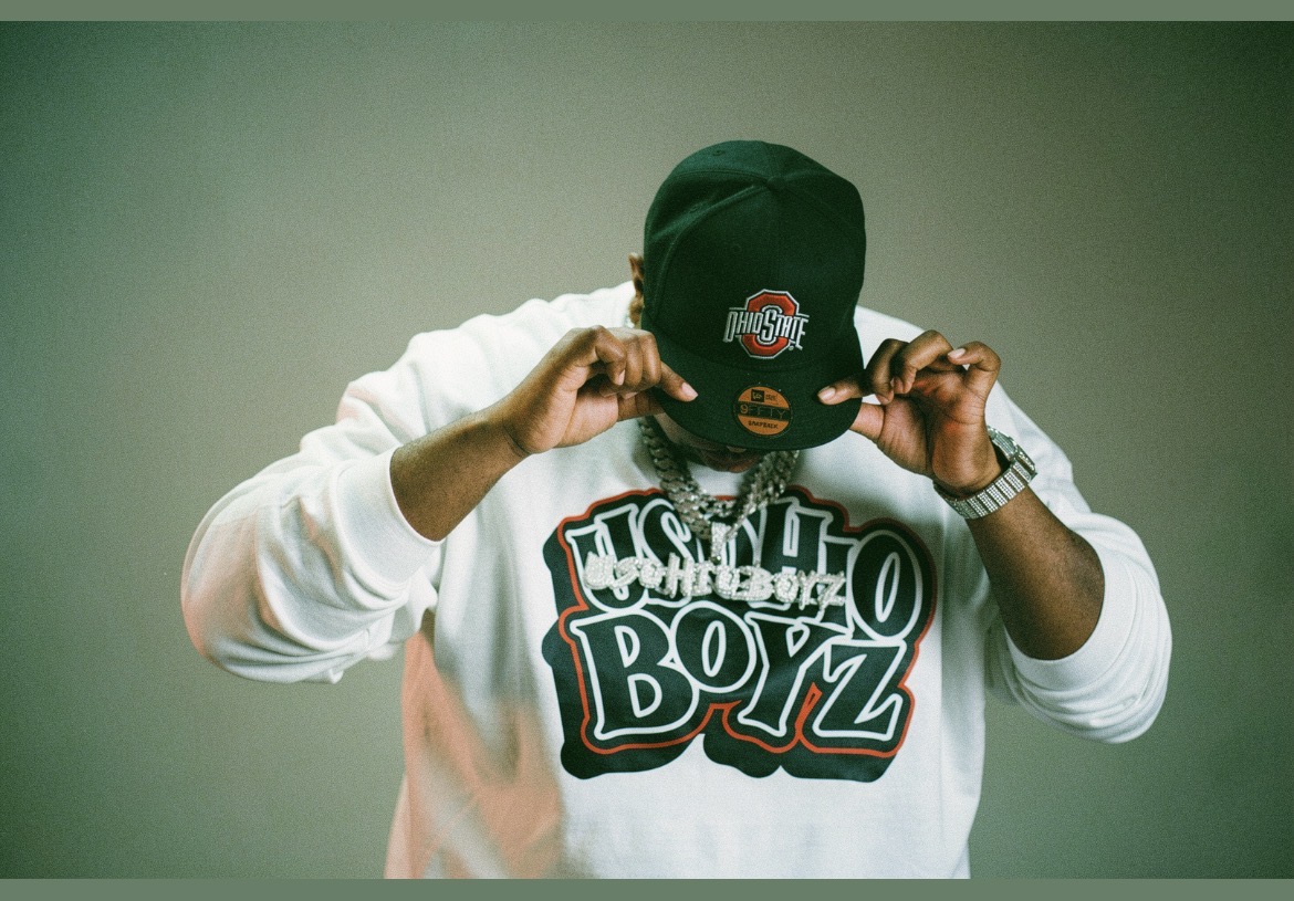 Person wearing a black cap and white sweatshirt, adjusting the cap, against a plain background.