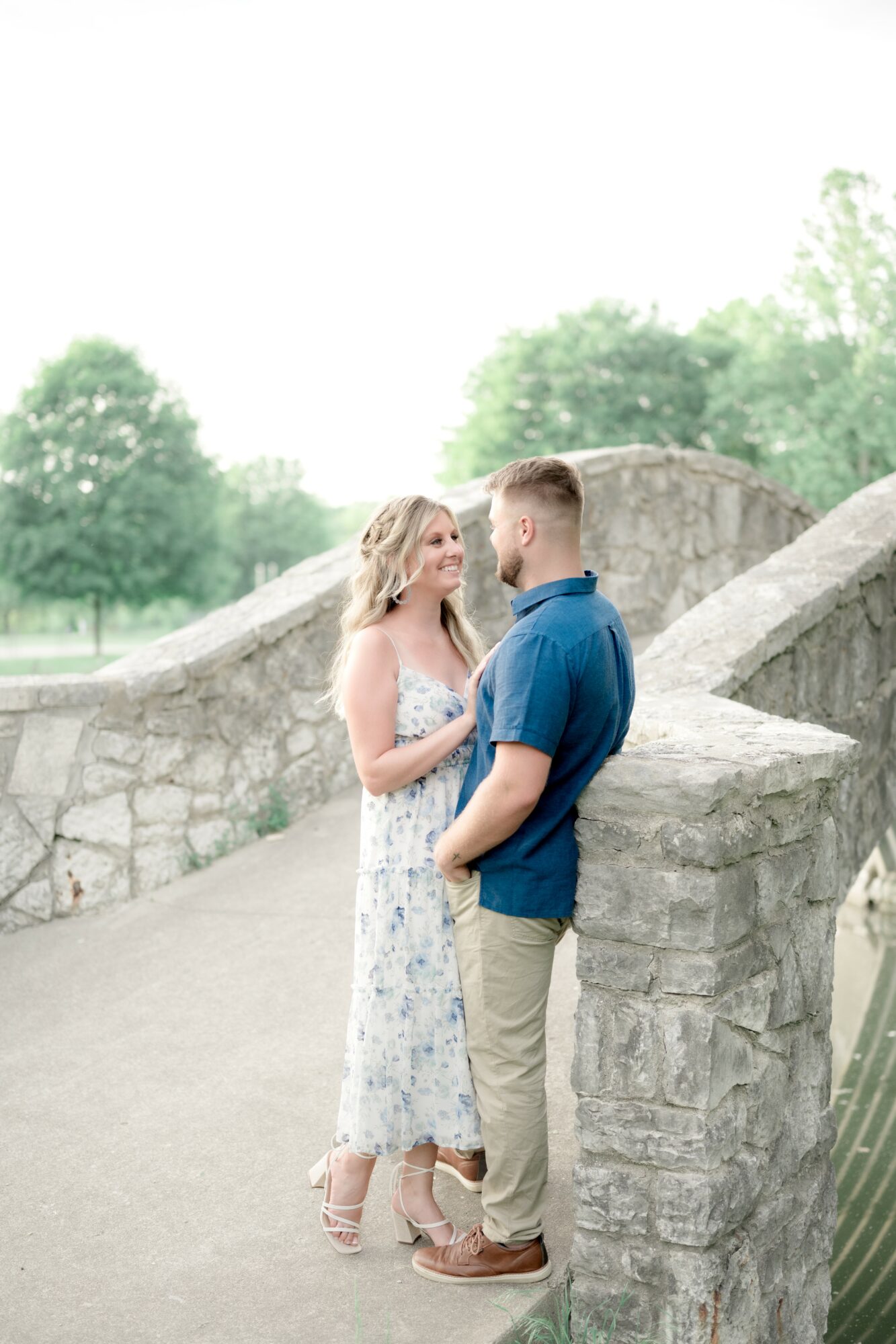 A couple stands close together outdoors near a stone wall, with trees in the background, engaging in conversation.