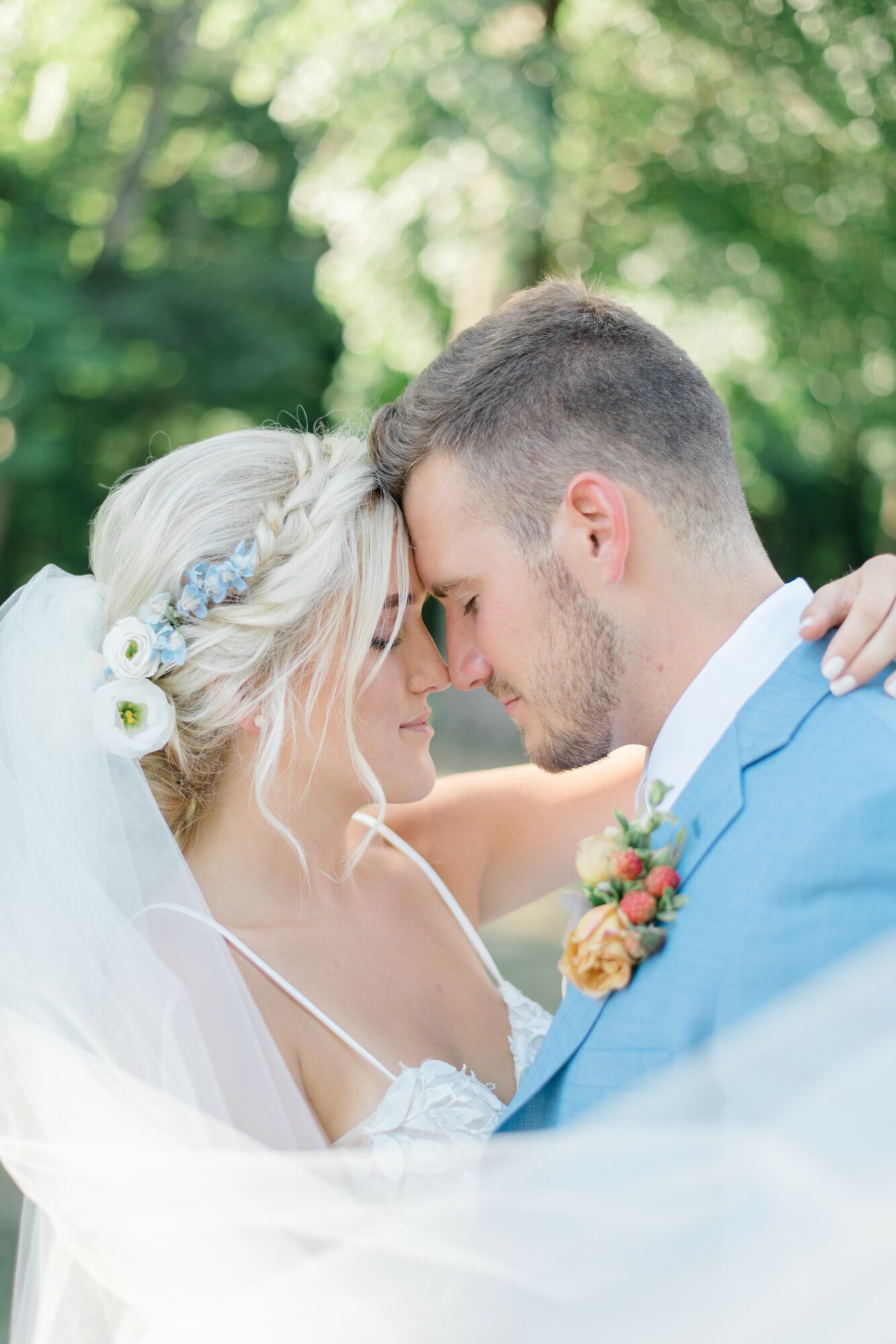 Bride and groom with foreheads touching outdoors, surrounded by greenery, bride wearing a floral headband, groom in a blue suit.