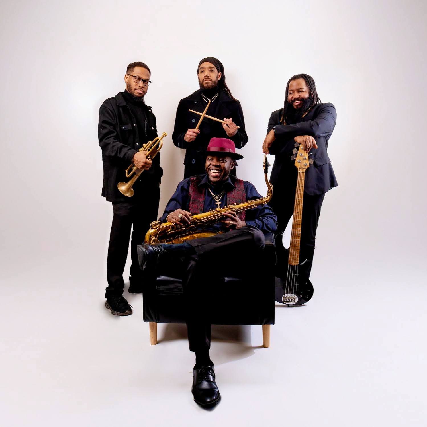 Four musicians with instruments pose against a plain white background. One seated with a saxophone, three standing with a trumpet, drumsticks, and bass guitar.
