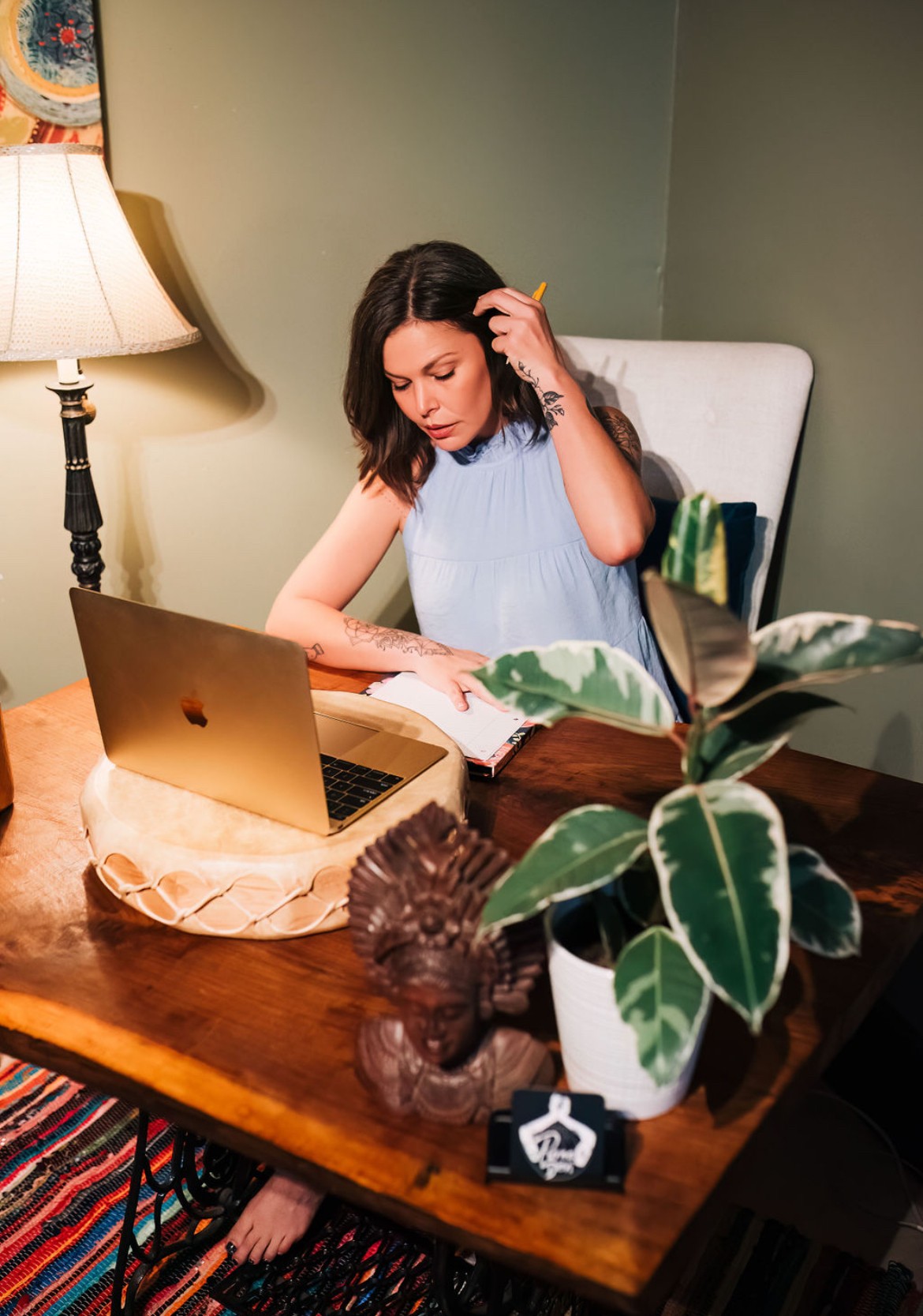 Woman sitting at a table, reading a book with a laptop open, next to a potted plant, in a cozy room.