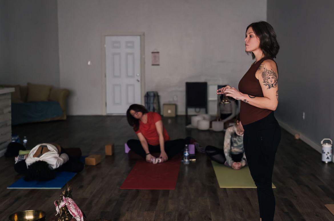 A woman with tattoos stands in a room with yoga mats, while a person practices yoga on a mat and another lies on the floor.