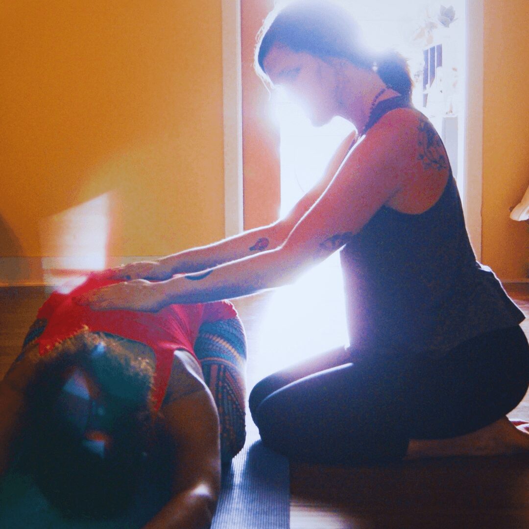 Person kneeling and stretching arms towards another person sitting on the floor, sunlight streaming through a window behind them.