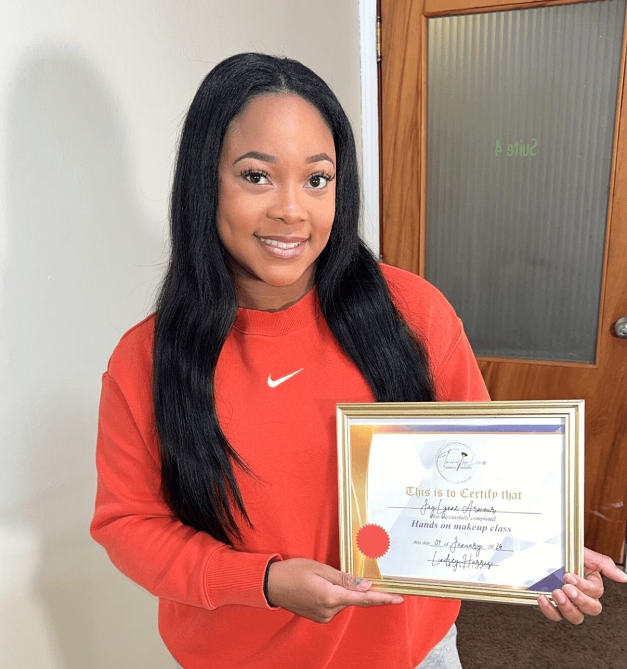 Young woman with long black hair holding a framed certificate, smiling, wearing a red Nike sweatshirt, standing indoors.
