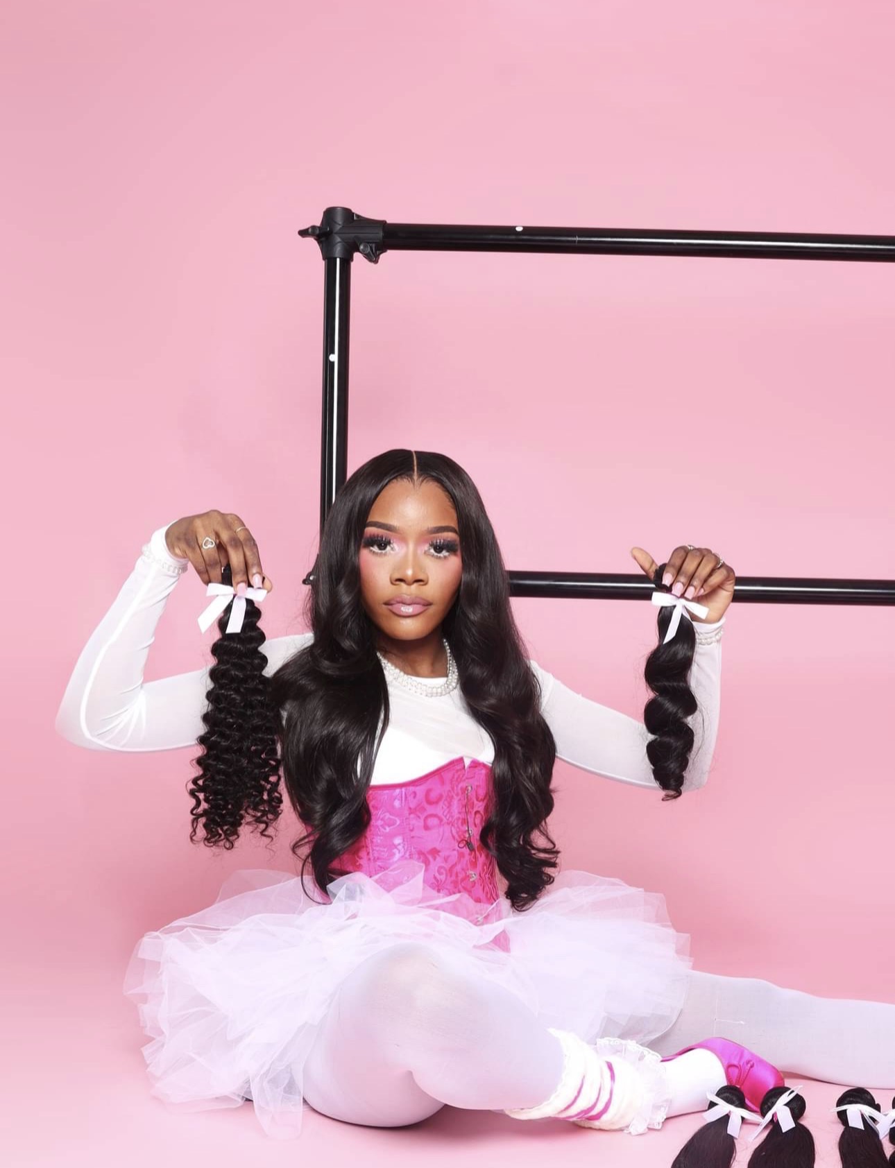Young woman with long black hair in a pink and white tutu, sitting on the floor against a pink background, holding black curly hair extensions.