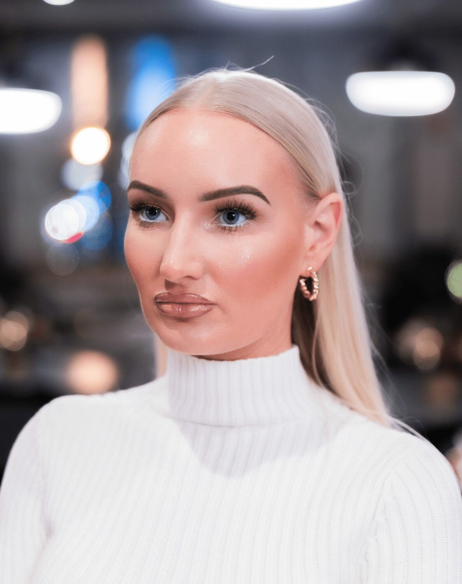 Woman with long blonde hair wearing a white turtleneck, earrings, and makeup, in an indoor setting with blurred background.