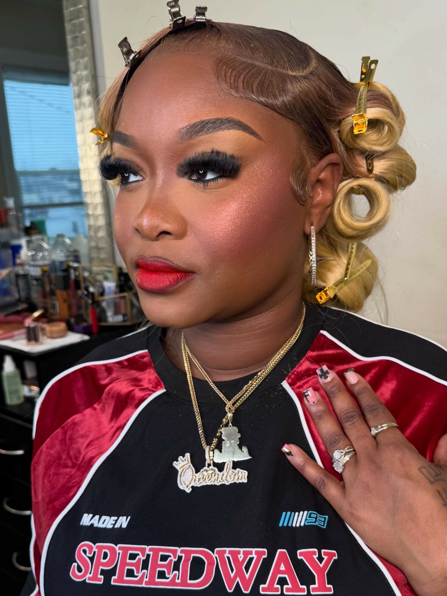 Close-up of a woman with styled hair, makeup, jewelry, and a racing jacket, in a room with makeup products.