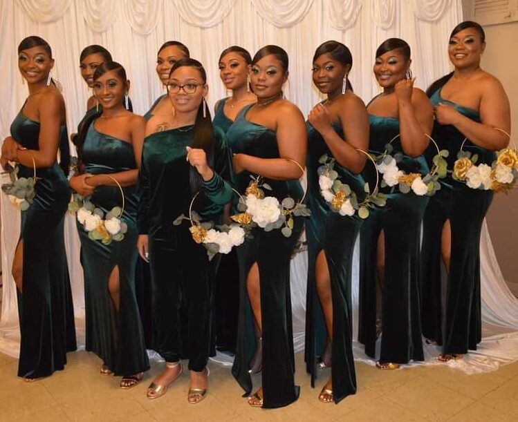 Group of nine women in matching dark green dresses holding white and yellow flowers, standing in front of a white curtain.