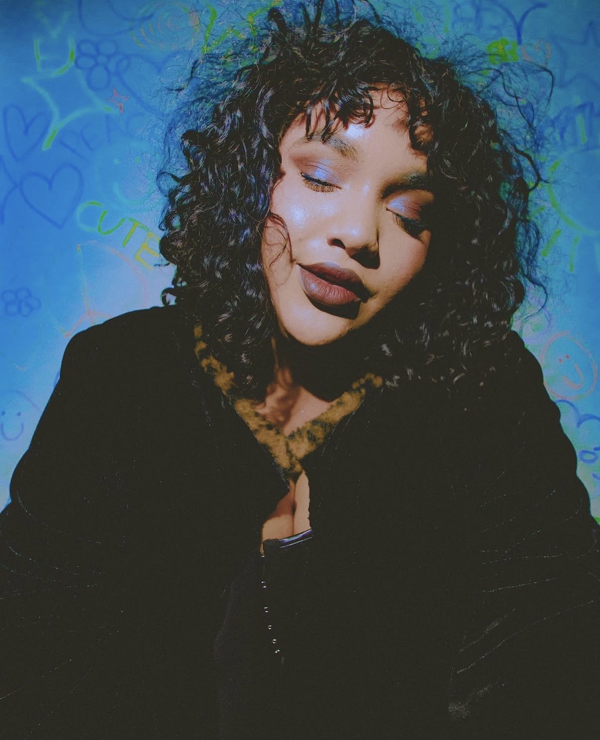 Woman with curly hair and closed eyes, wearing dark clothing, against a blue background with patterns.