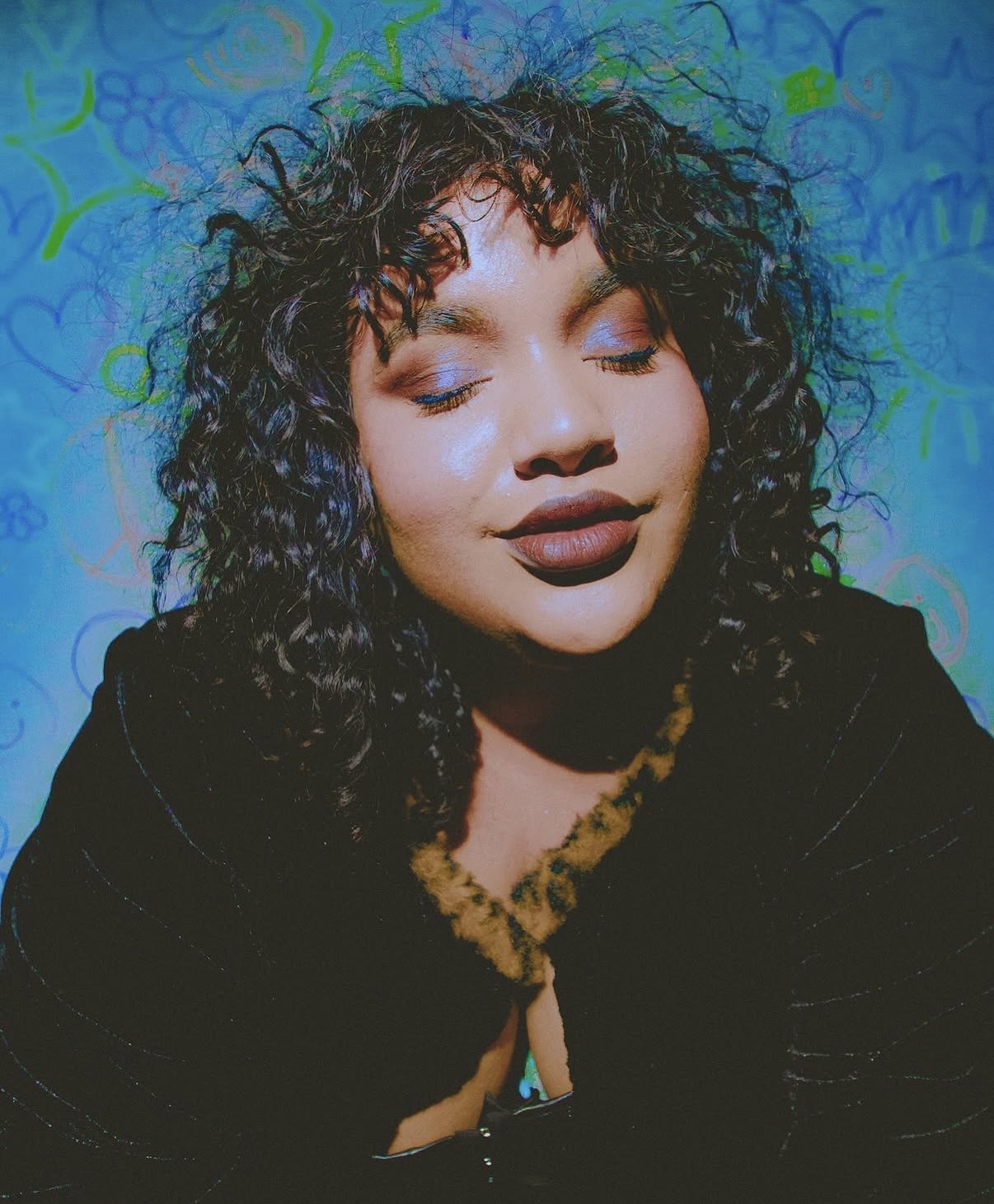 Young woman with curly hair and closed eyes, smiling, against a colorful background.