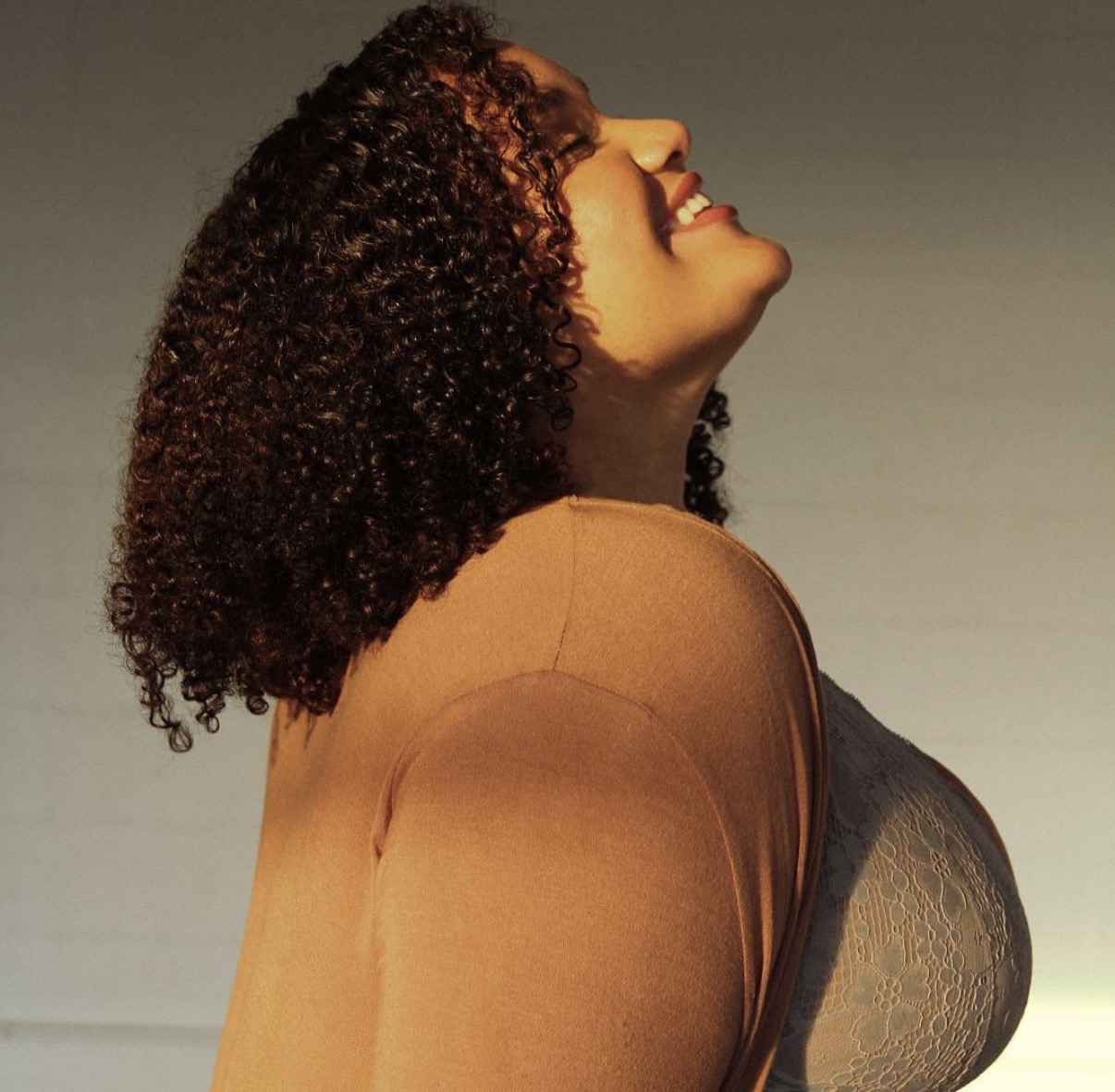Woman with curly hair smiling and looking upward, wearing a brown top and gray bra, against a plain background.