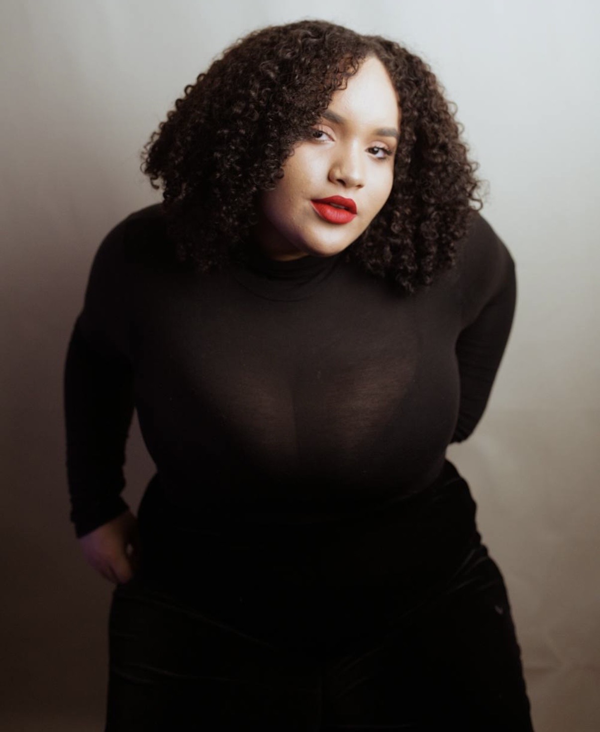Woman with curly hair and red lipstick wearing a black top, standing against a plain background.