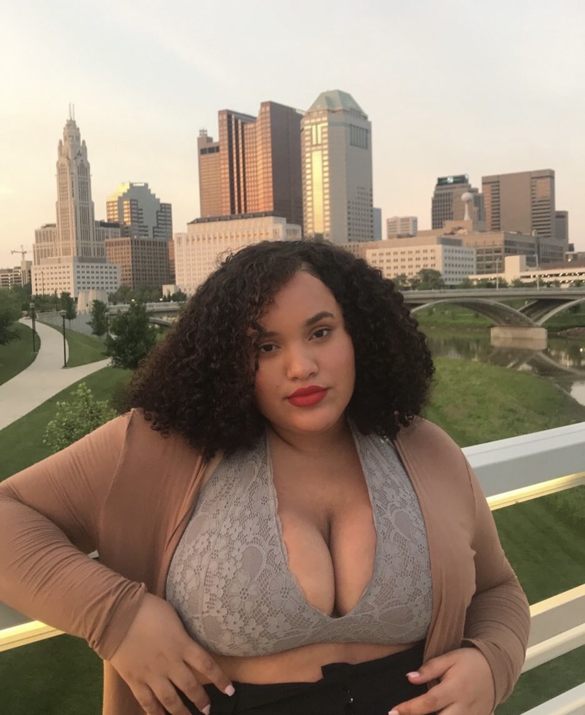 Woman with curly hair and red lipstick standing outdoors with city skyline in background.