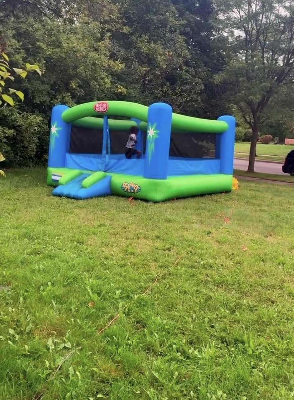 Inflatable bounce house with blue, green, and yellow colors set up on a grassy area, surrounded by trees and a park path.