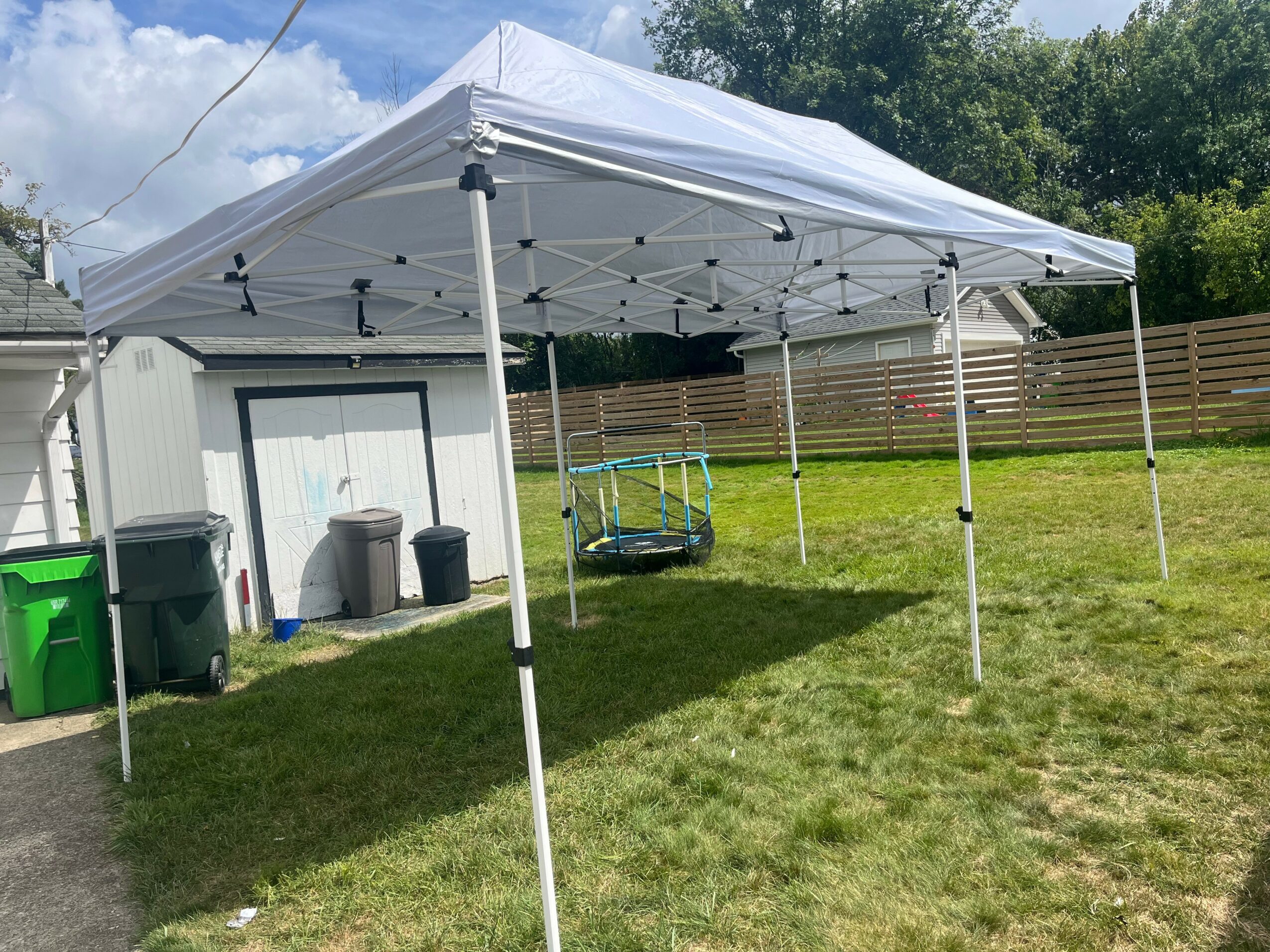 White canopy tent set up on a grassy yard with a shed, trash cans, and a wooden fence in the background.