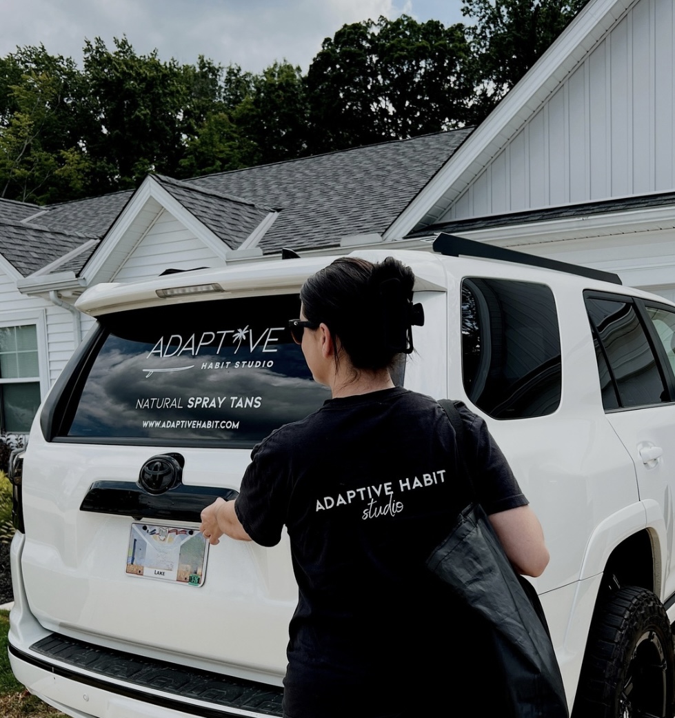 Person with short dark hair wearing a black t-shirt and carrying a black bag stands near a white vehicle with advertising on the rear window, in front of a house with a gray roof and trees.