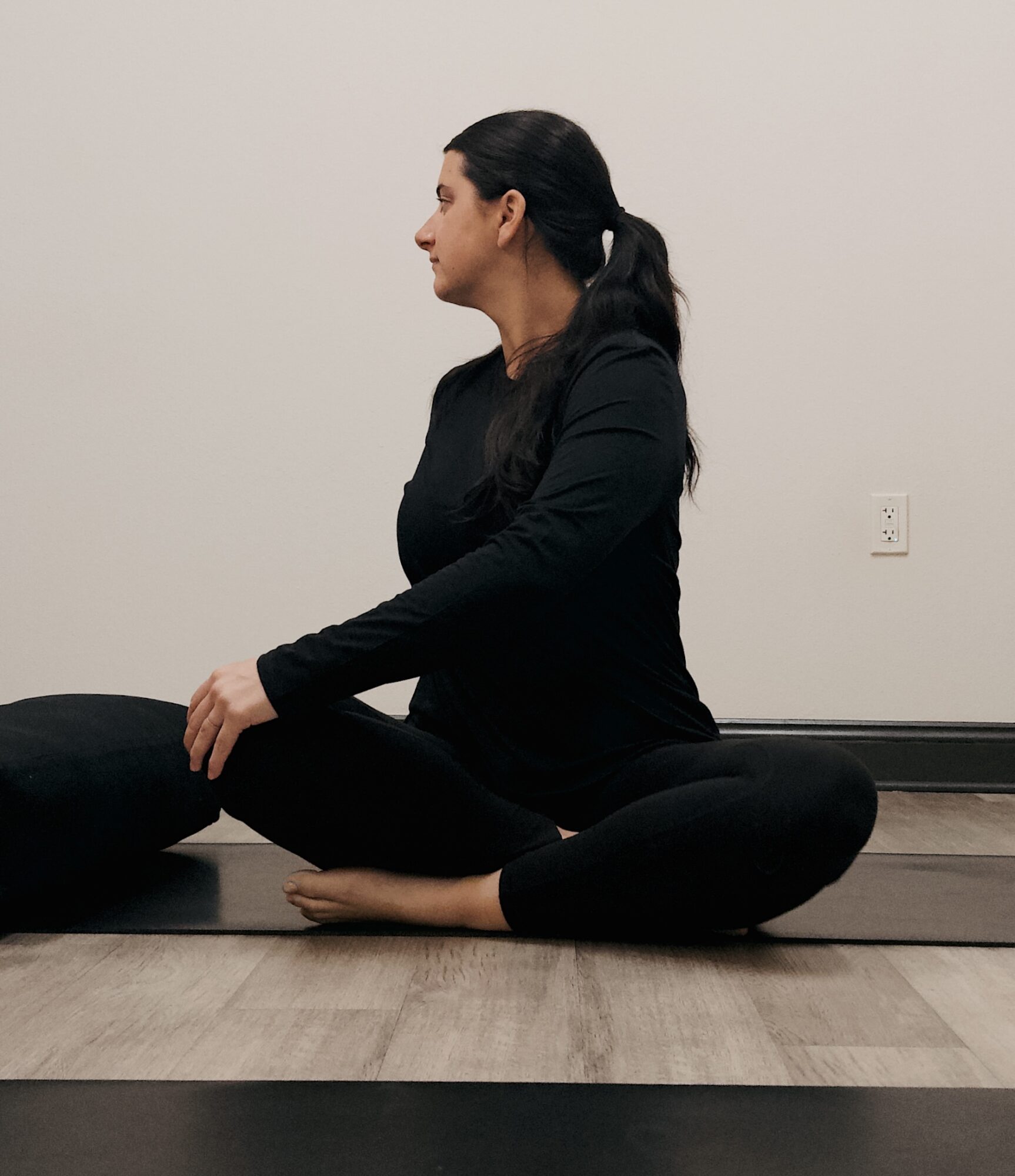 Woman practicing yoga seated cross-legged on a mat, facing left, in a room with plain wall and electrical outlet.