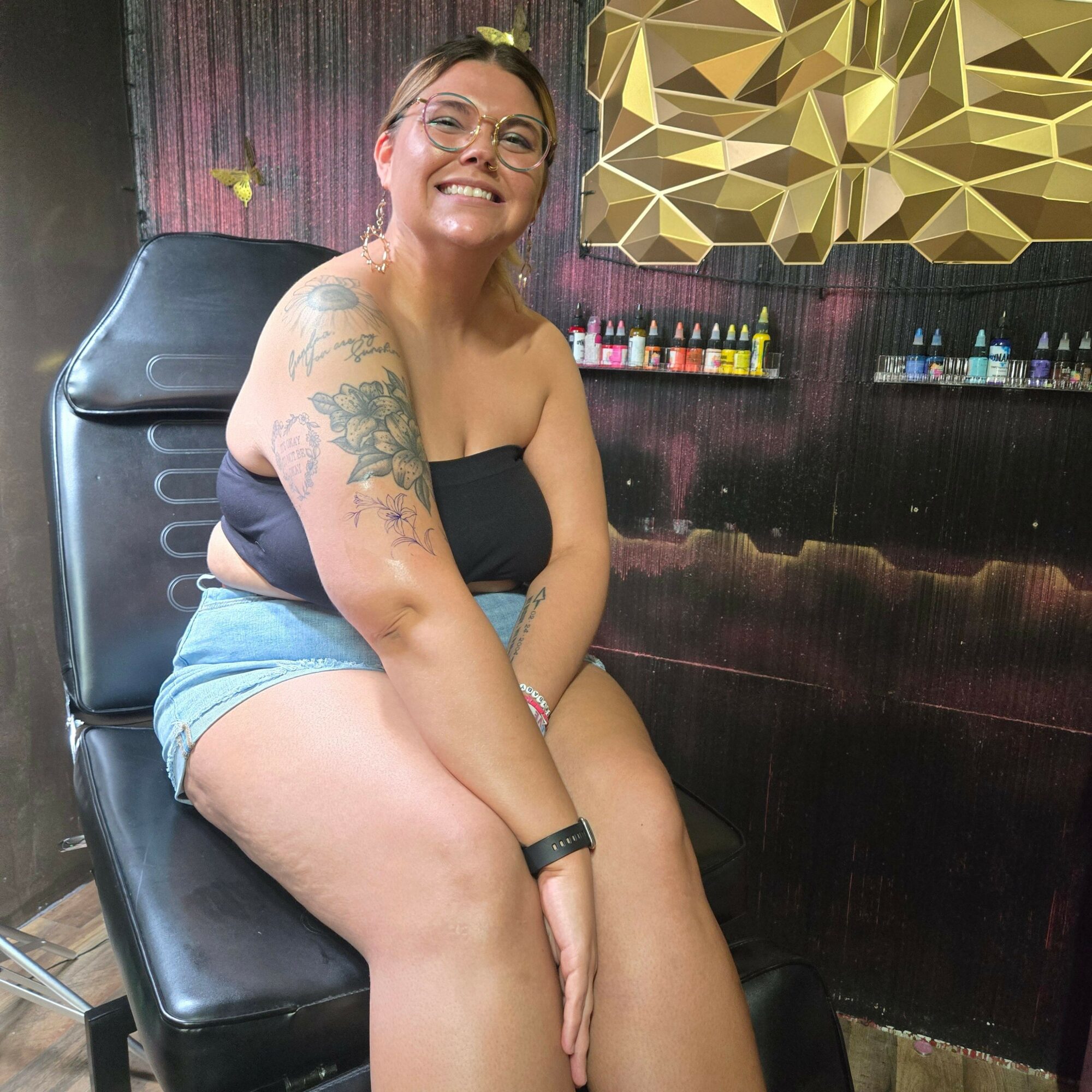 Smiling woman with glasses and tattoos, sitting on a black bench in a room with geometric wall decor and shelves.