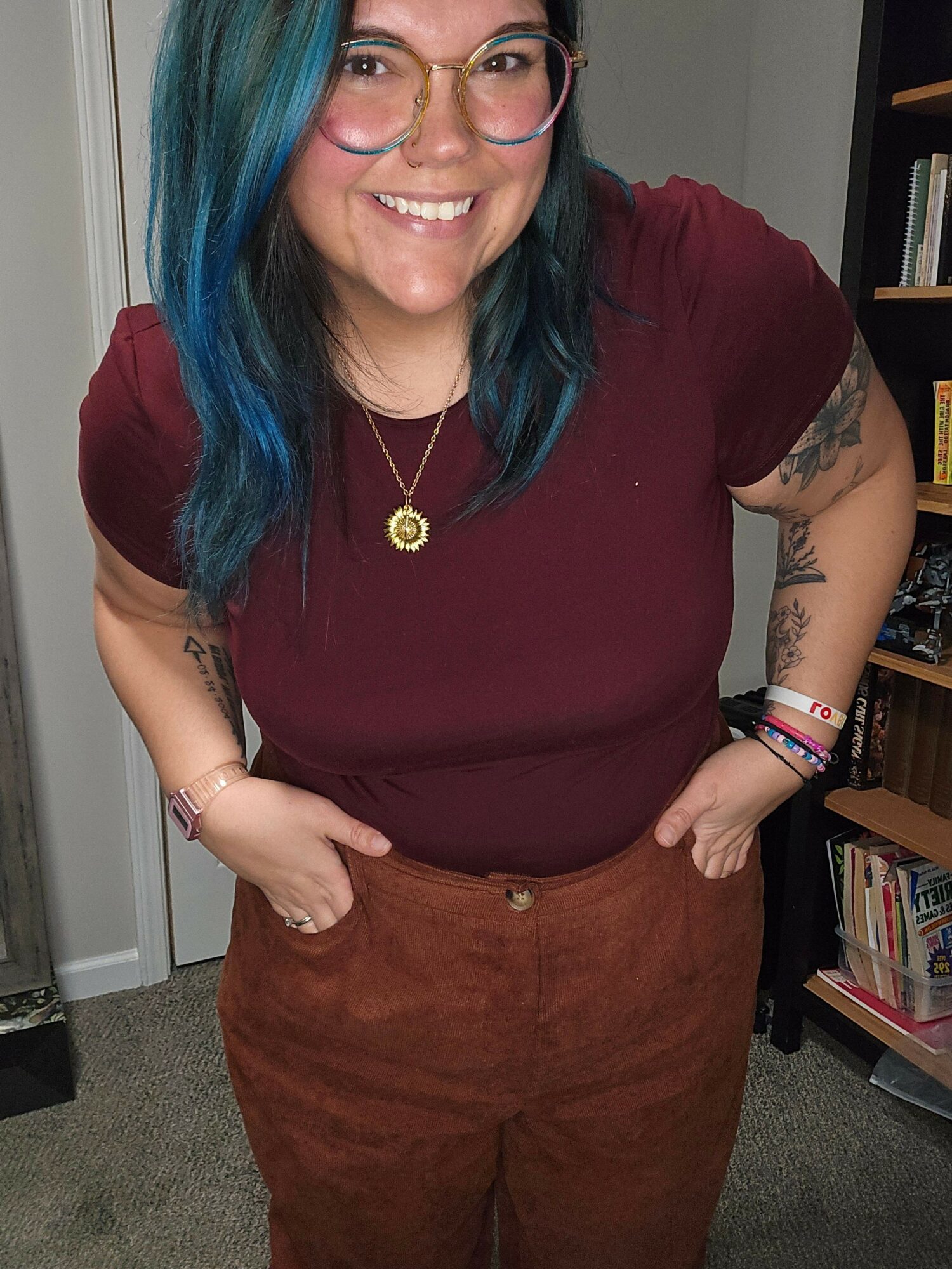 Woman with blue hair, glasses, and tattoos smiling, wearing a maroon top and brown pants, standing indoors.