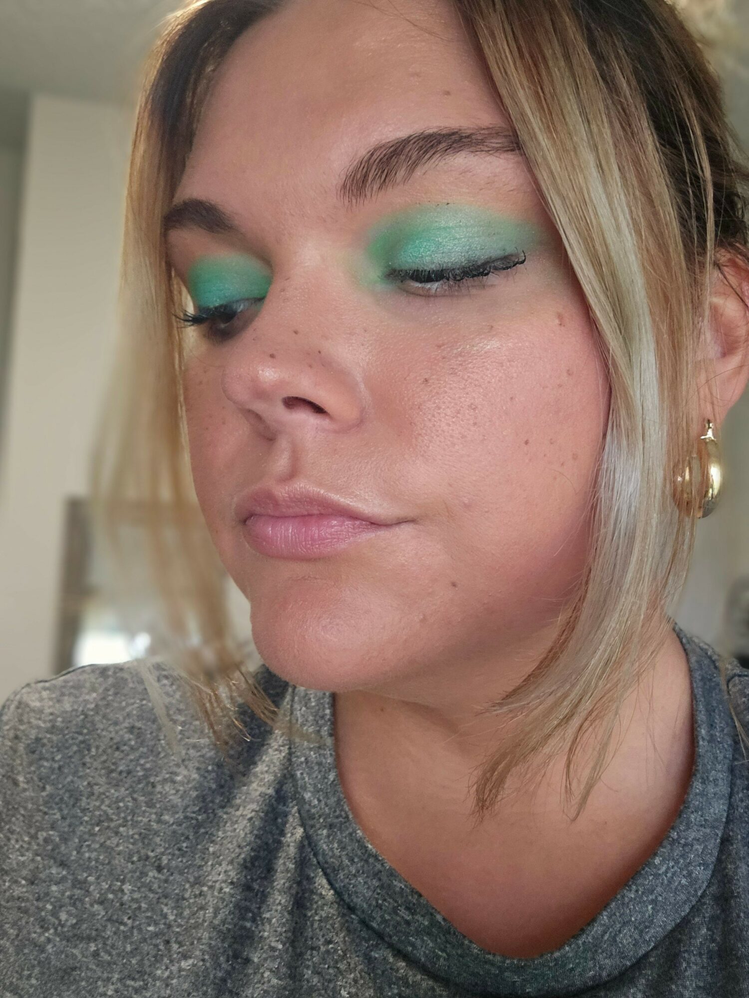 Close-up of a woman with closed eyes, wearing bright green eyeshadow and a gray top.
