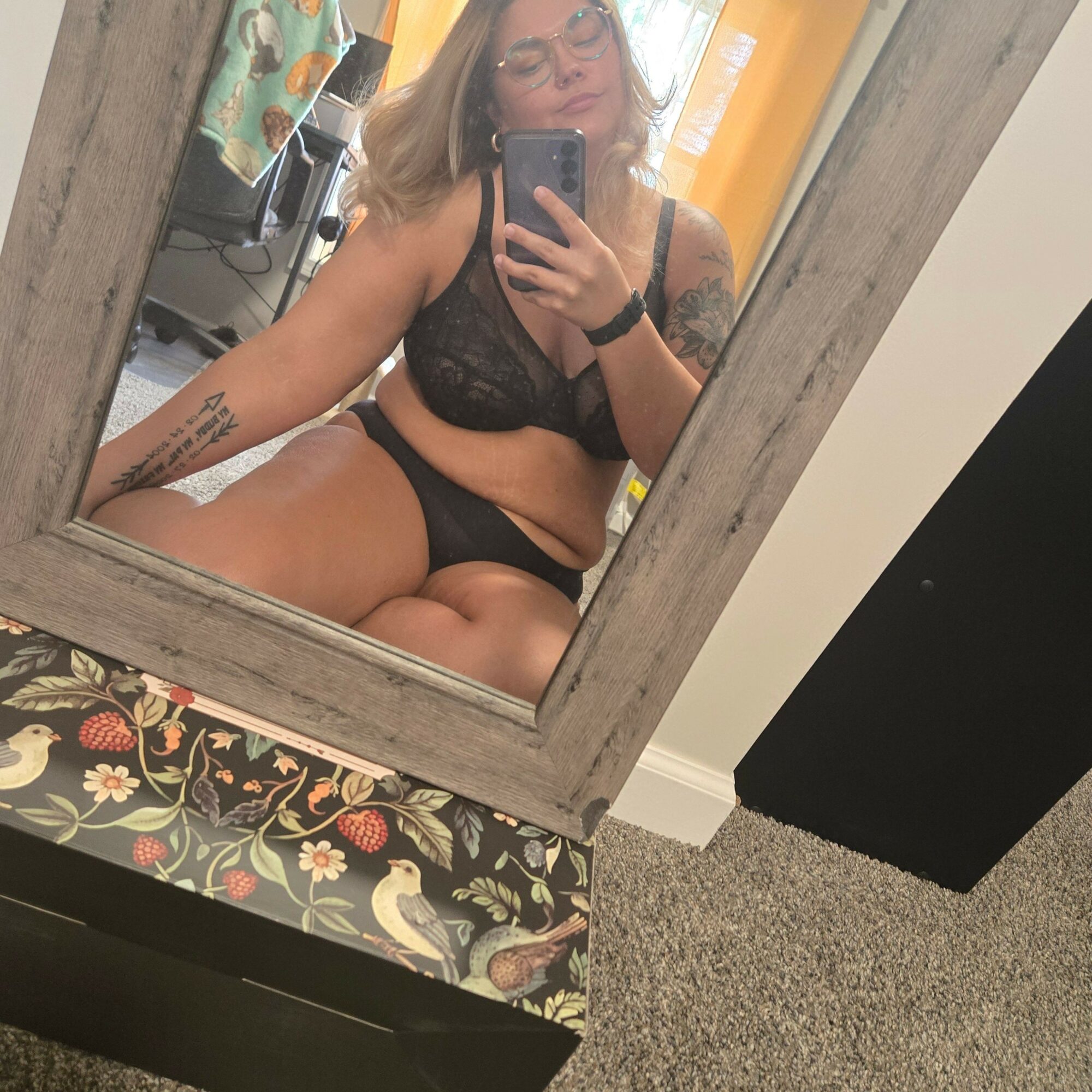 Person taking a mirror selfie in a room, wearing black lingerie, with a tattoo on their arm, and holding a phone.