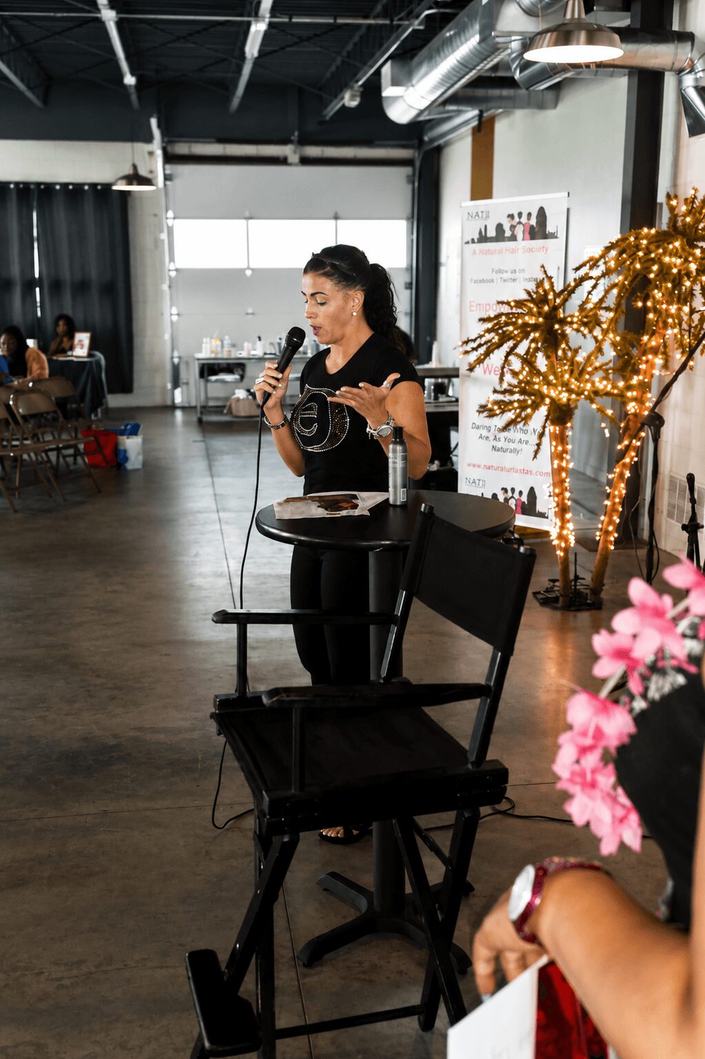 Woman speaking into microphone in industrial-style room with black ceiling, large windows, and decorative lights, holding a paper.