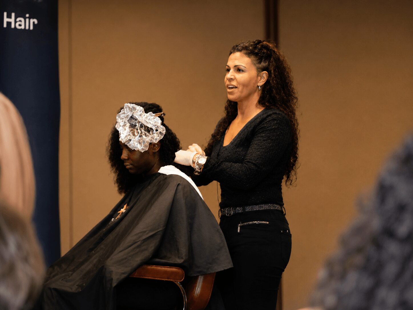 A woman with curly hair stands next to a seated person with a large white hair accessory, in a salon setting.
