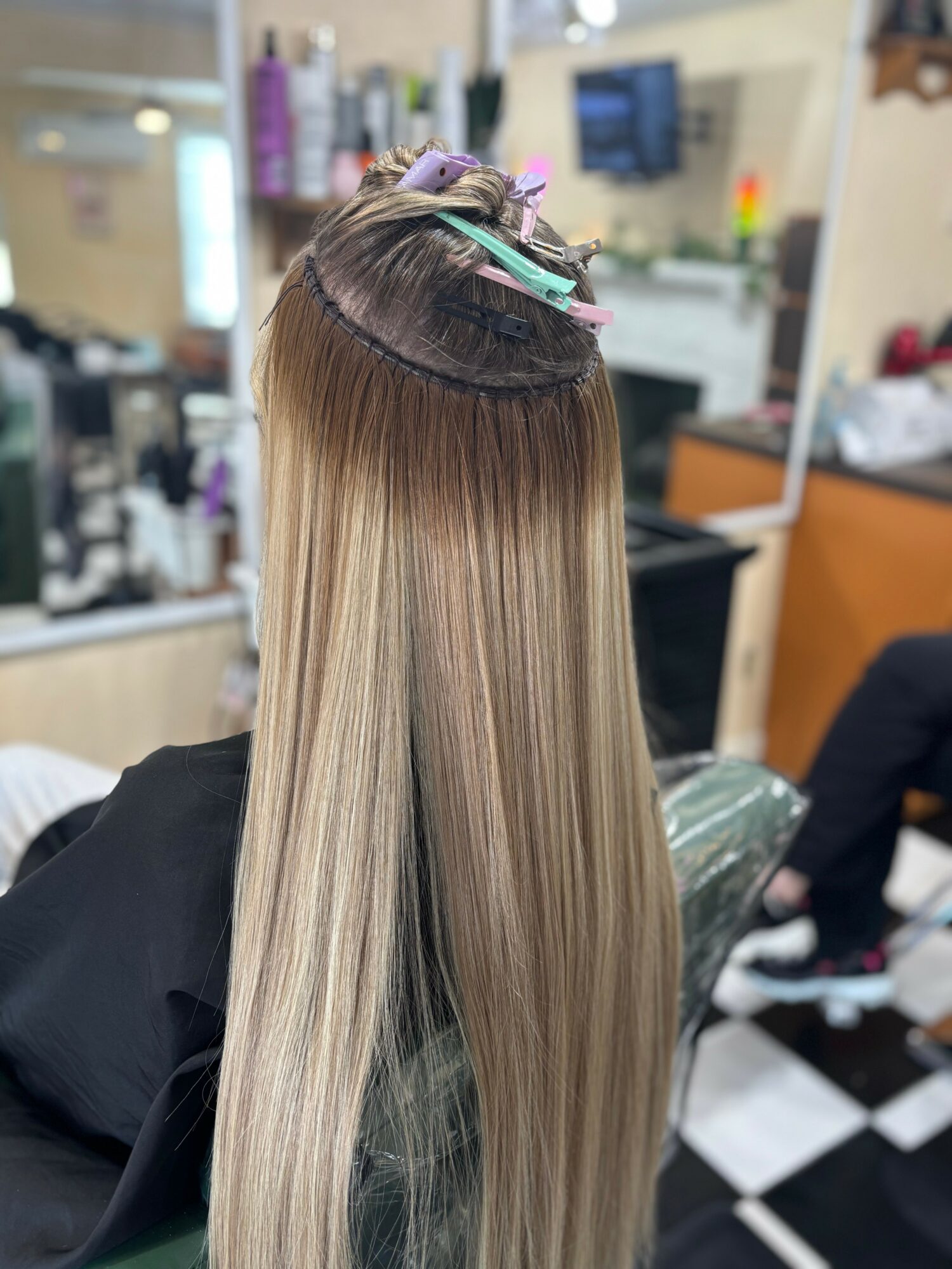 Back of a person with long, straight, blonde hair in a salon, hair sectioned and clipped, with salon equipment in background.