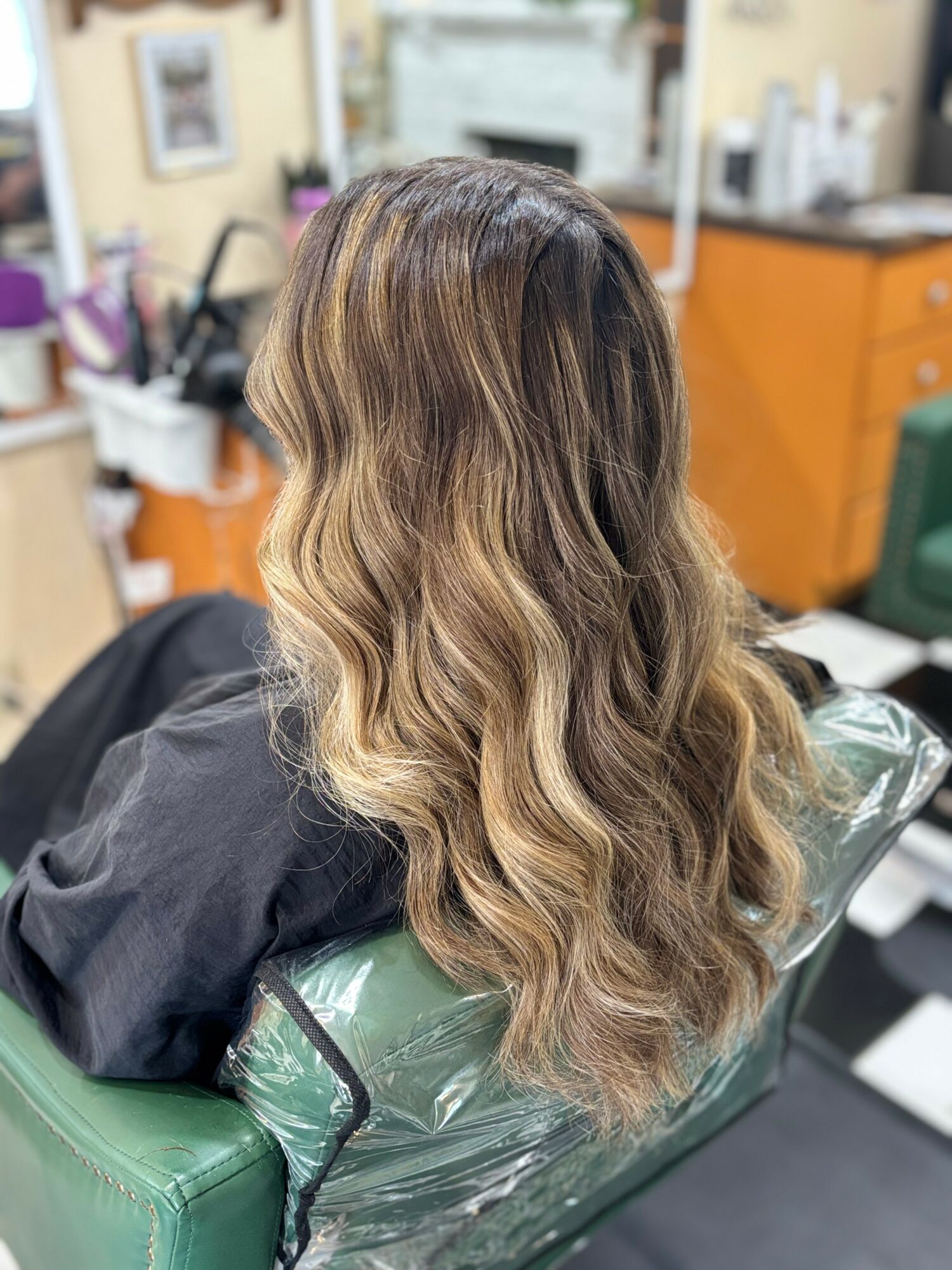 Person with long, wavy hair sitting in a salon chair, facing away, with salon equipment in the background.