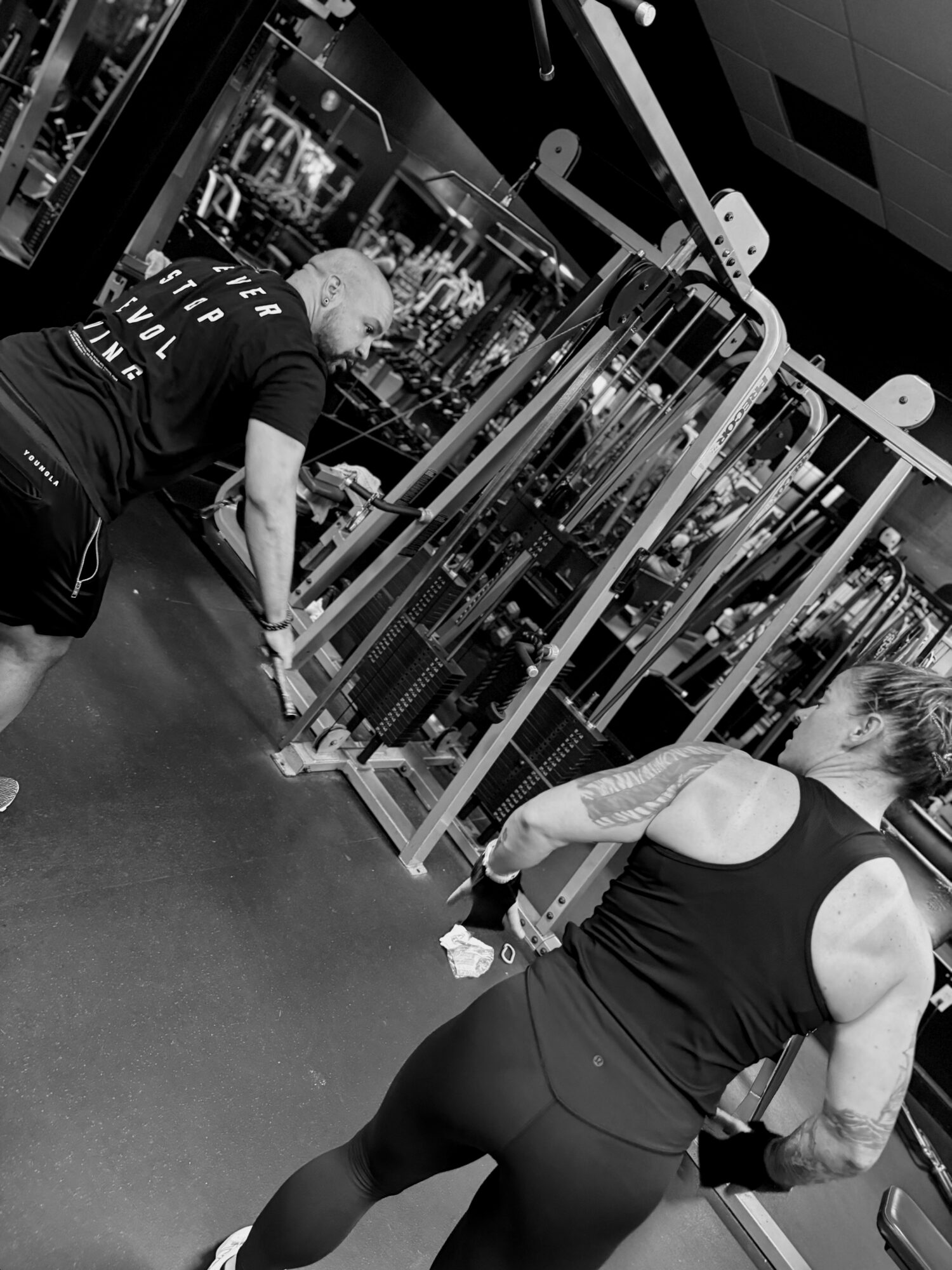 Two women in a gym, one adjusting equipment and the other standing with her back to the camera, in black workout clothes.