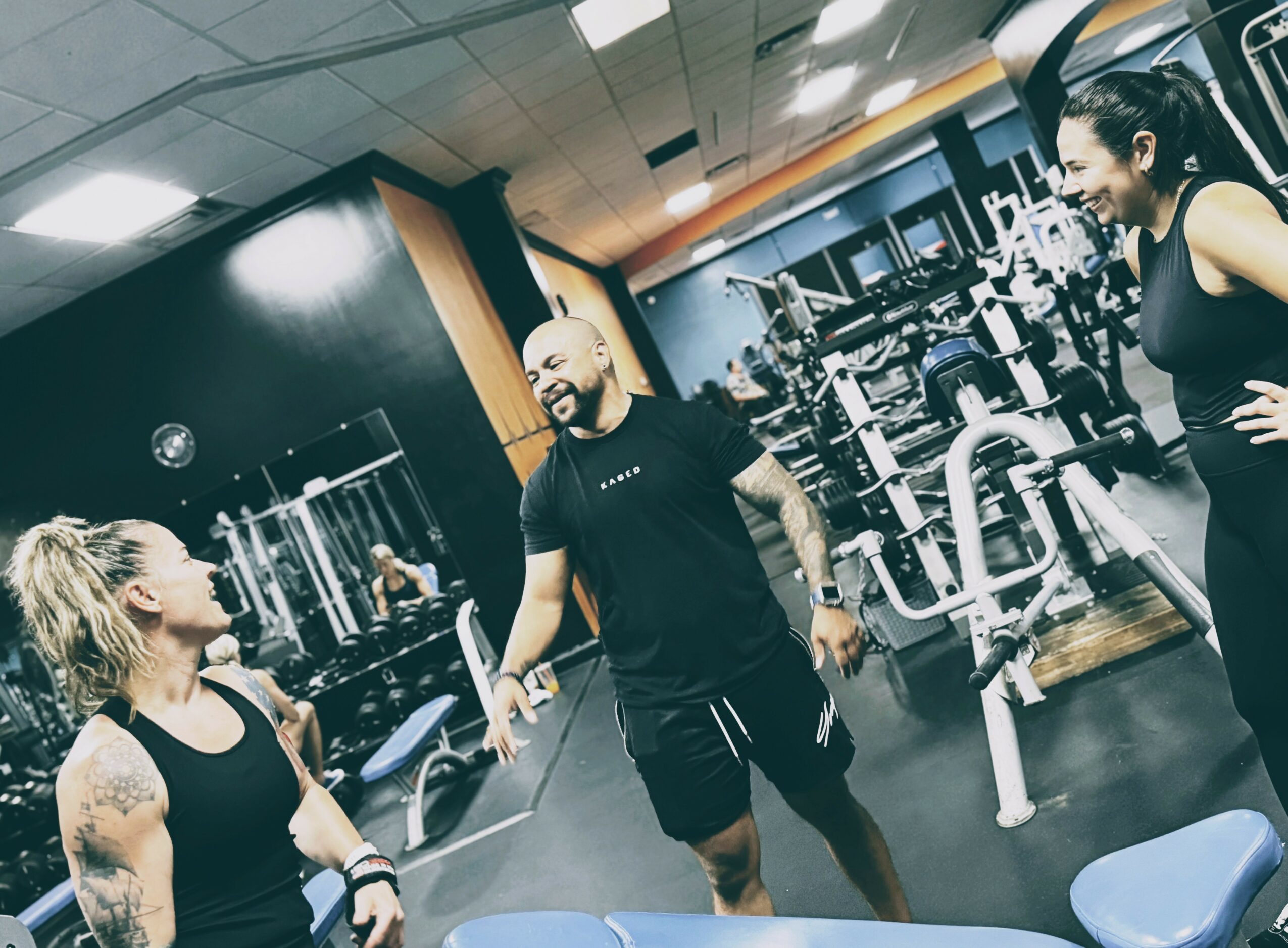 Three people in a gym, two women and one man, engaged in conversation near workout equipment.