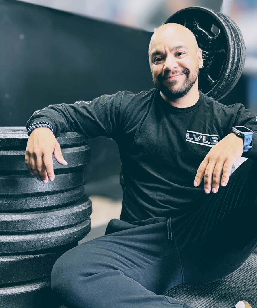 Man sitting on the floor next to stacked weight plates, smiling, in a gym setting.