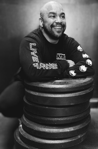 Smiling man with a beard and shaved head sitting at a table with stacked plates, wearing a long-sleeve shirt with text and graphics.