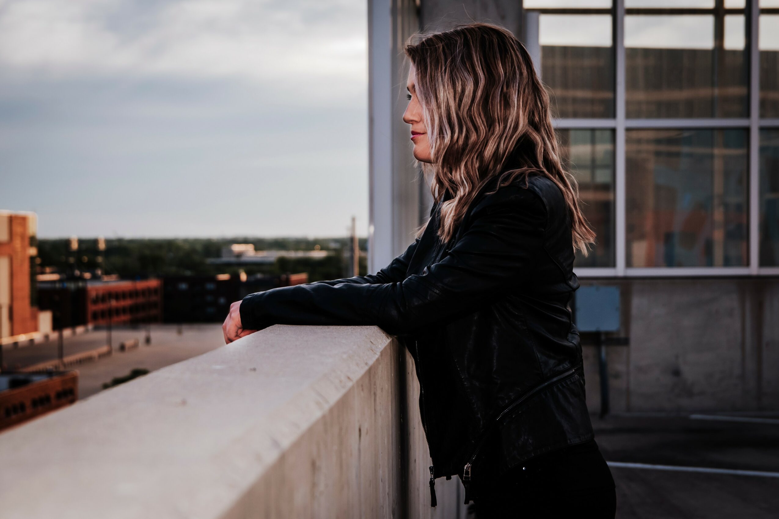 Woman with wavy hair leaning on a rooftop ledge, cityscape in background, wearing a black jacket.