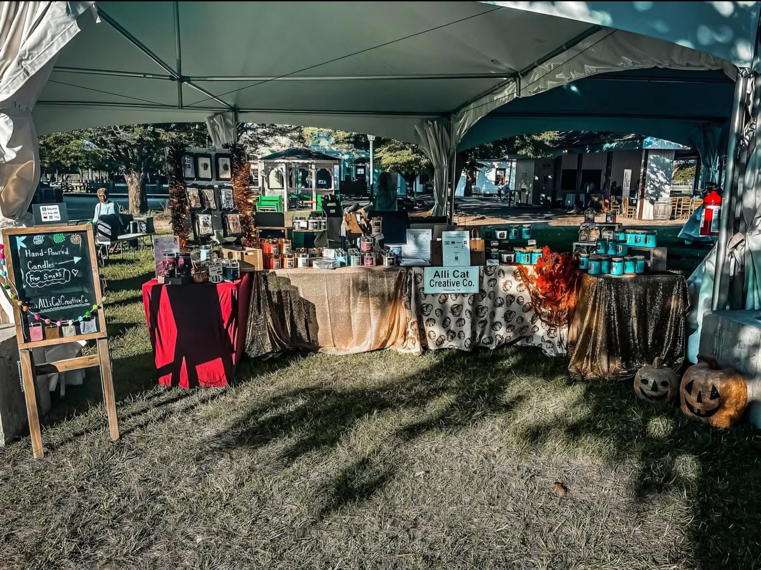 Outdoor market stall with tables displaying various items under a tent, with a grassy area in front.