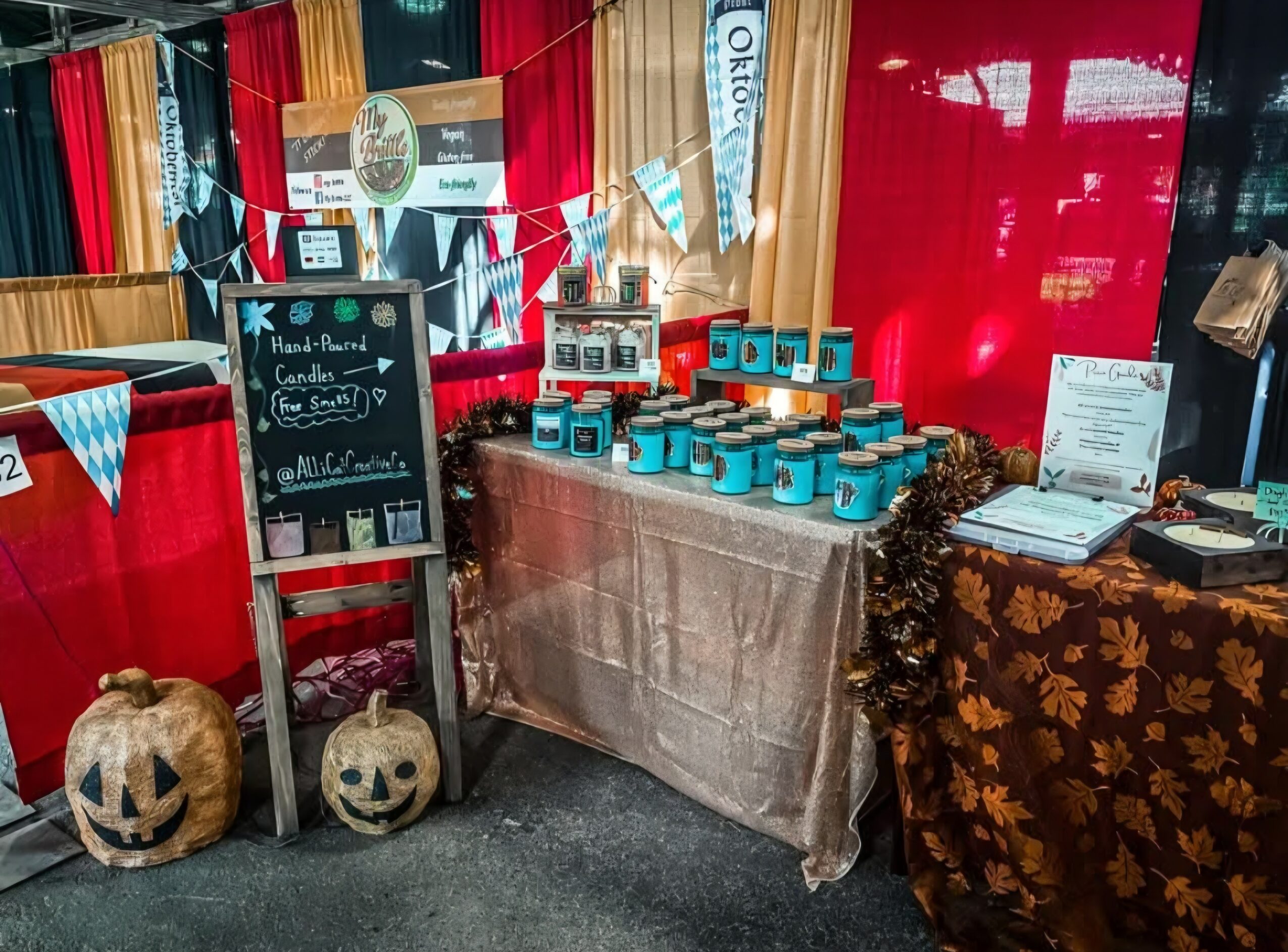 Display table with blue jars, blackboard sign, and pumpkins, decorated with bunting and autumn-themed cloths, at an indoor event.