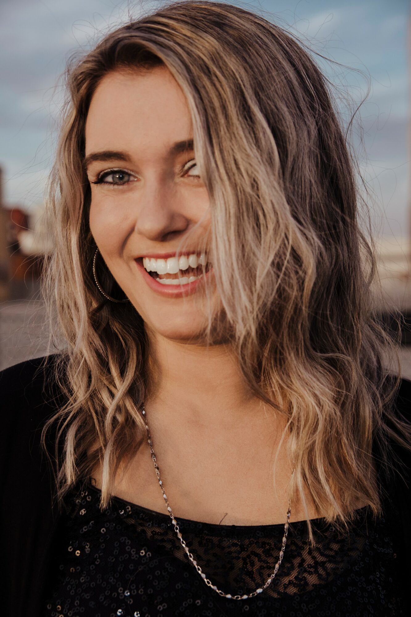 Young woman with wavy hair smiling outdoors, wearing a black top and a necklace.
