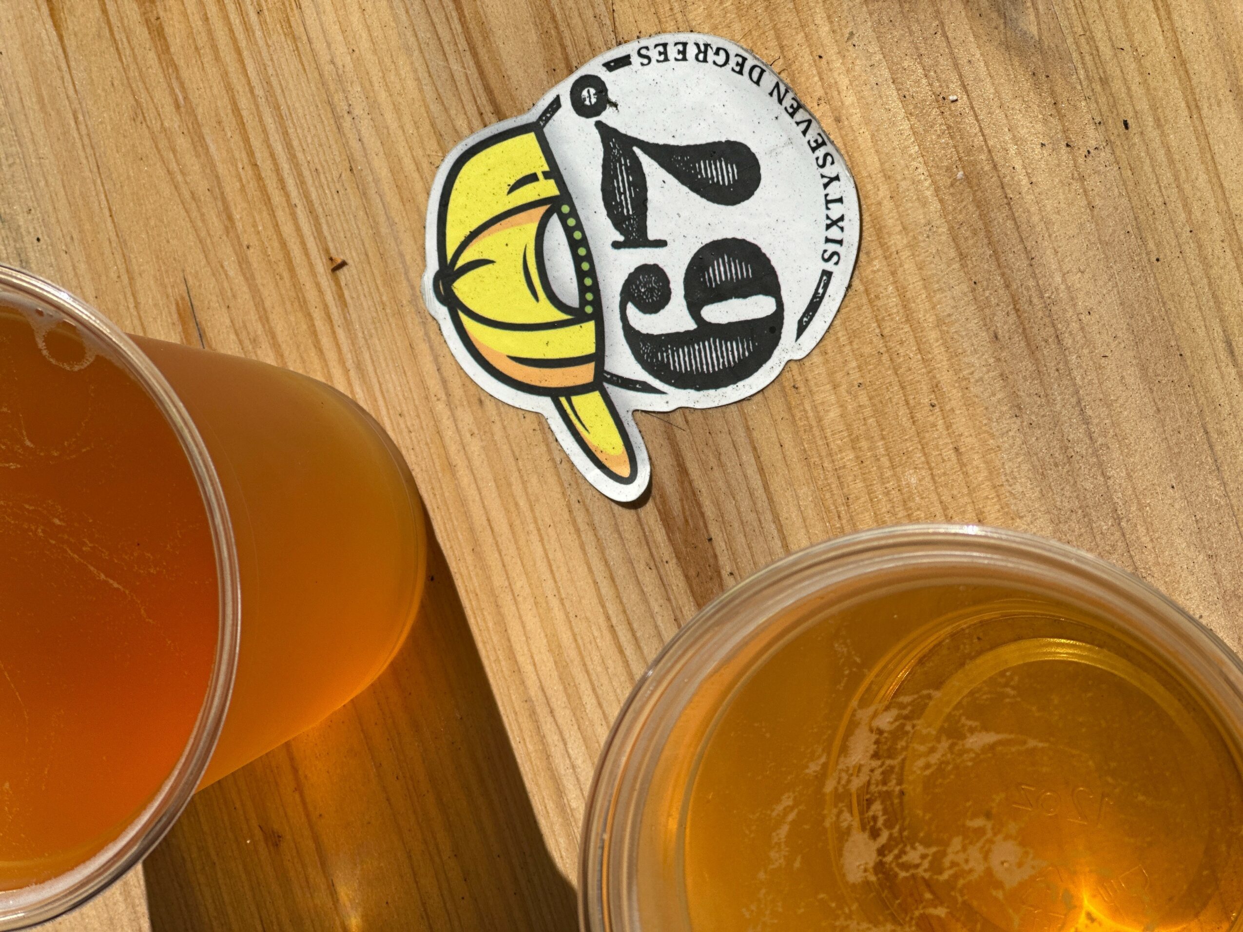 A glass of beer, a sticker with a cartoon character wearing a yellow hat, and a wooden surface.
