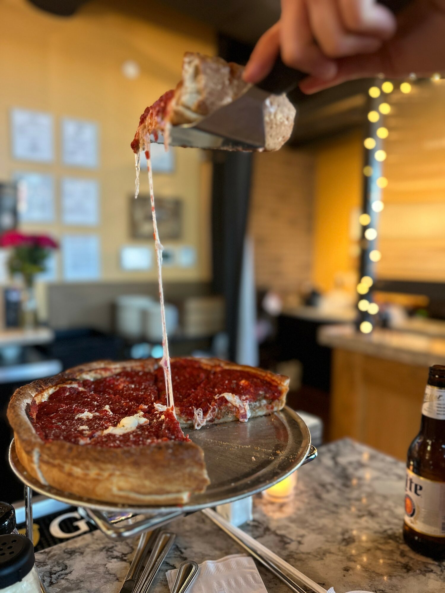 Person lifting a slice of pizza with cheese stretching, on a silver tray in a cozy restaurant setting.