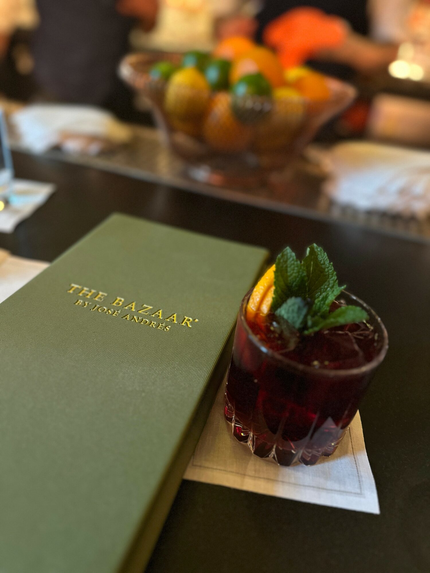 A dark red cocktail garnished with mint and citrus slices, placed on a white napkin next to a green menu, in a dimly lit setting.
