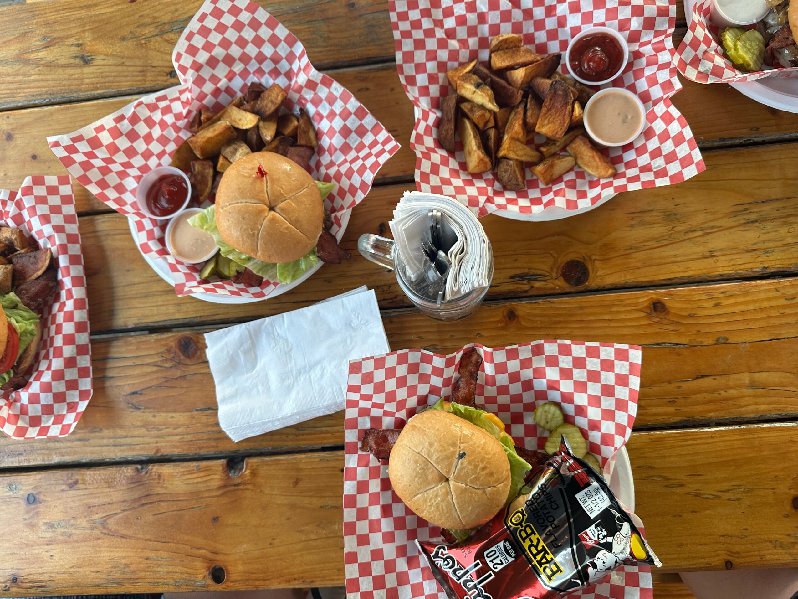Three baskets of food with sandwiches, potato wedges, and dipping sauces on a wooden table, with a napkin and utensils in the center.