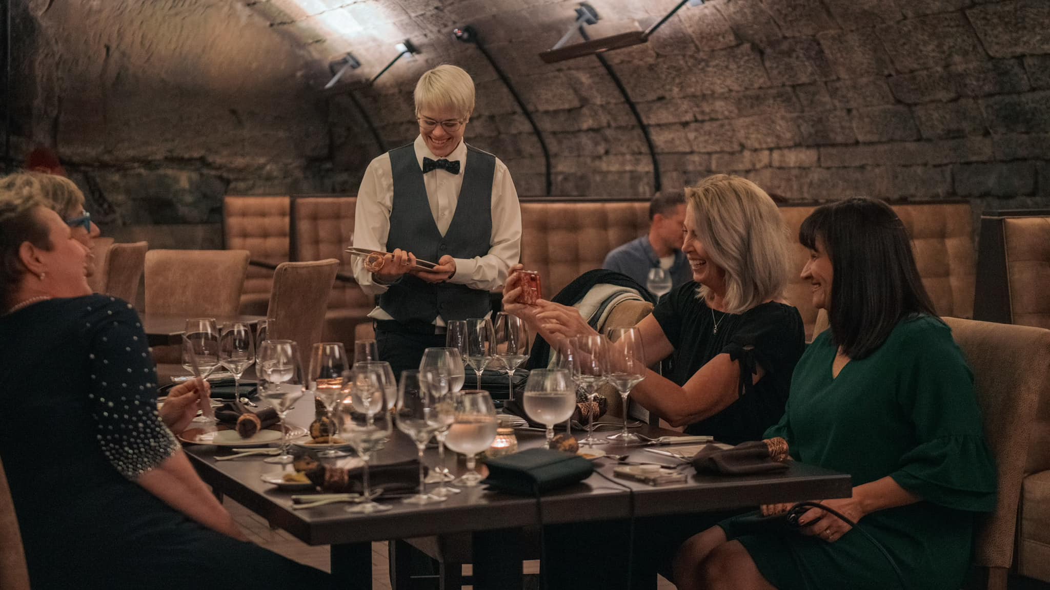 People dining at a restaurant table with a server standing nearby, in a dimly lit setting.